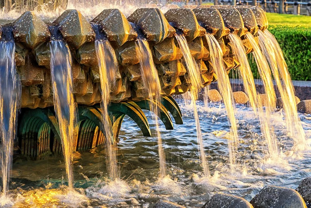 A stone fountain with multiple water streams flowing into a pond, illuminated by sunlight. Charleston's Waterfront Park. Pineapple Fountain. Photography Classes in Charleston SC. Camera Classes in Charleston SC
