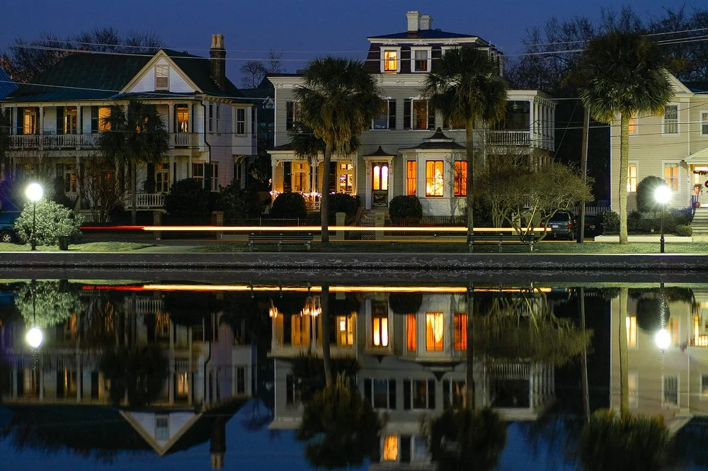 Nighttime view of three Victorian-style houses with porch lights on, reflected in a calm body of water in the foreground, with palm trees and streetlights along the sidewalk. Photography Classes in Charleston SC. Camera Classes in Charleston SC