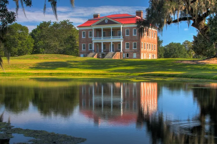 A large, historic brick mansion with a red roof, situated behind a pond reflecting the house and trees, in a lush green landscape. Drayton Hall. Photography Classes in Charleston SC. Camera Classes in Charleston SC