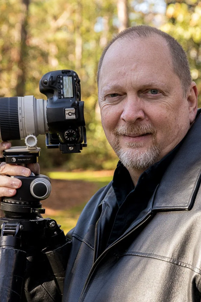The Owner holding a camera on a tripod outdoors with blurred trees in the background.