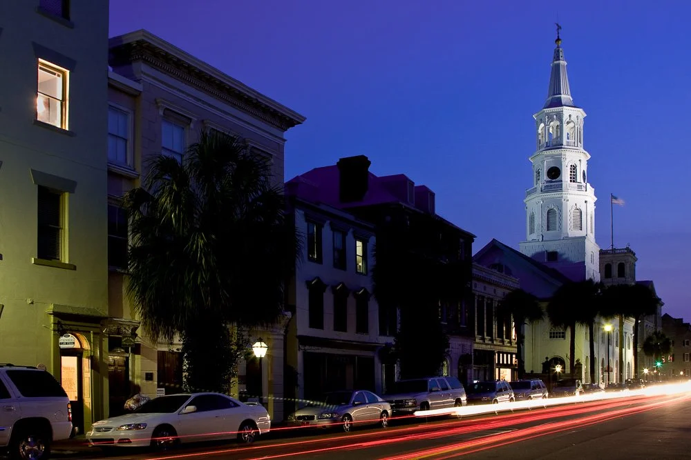 Street view of historic buildings with a church steeple in Charleston, South Carolina during dusk, with cars parked along the street and light trails from moving vehicles. Photography Classes in Charleston SC. Camera Classes in Charleston SC