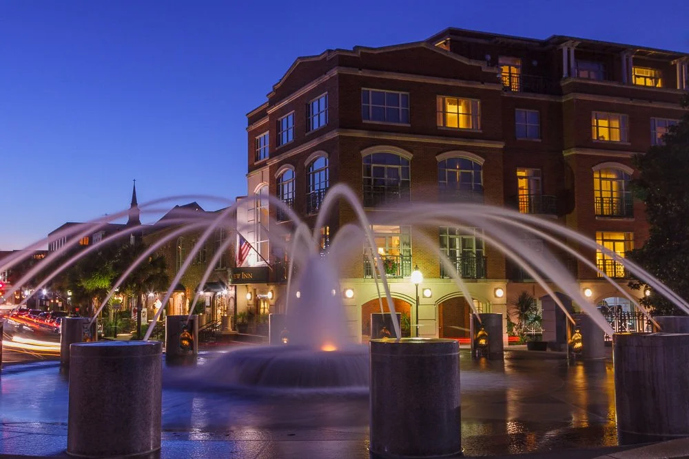 Night view of a lit fountain with water jets in front of a brick building with lit windows and a church steeple in the background. Photography Classes in Charleston SC. Camera Classes in Charleston SC