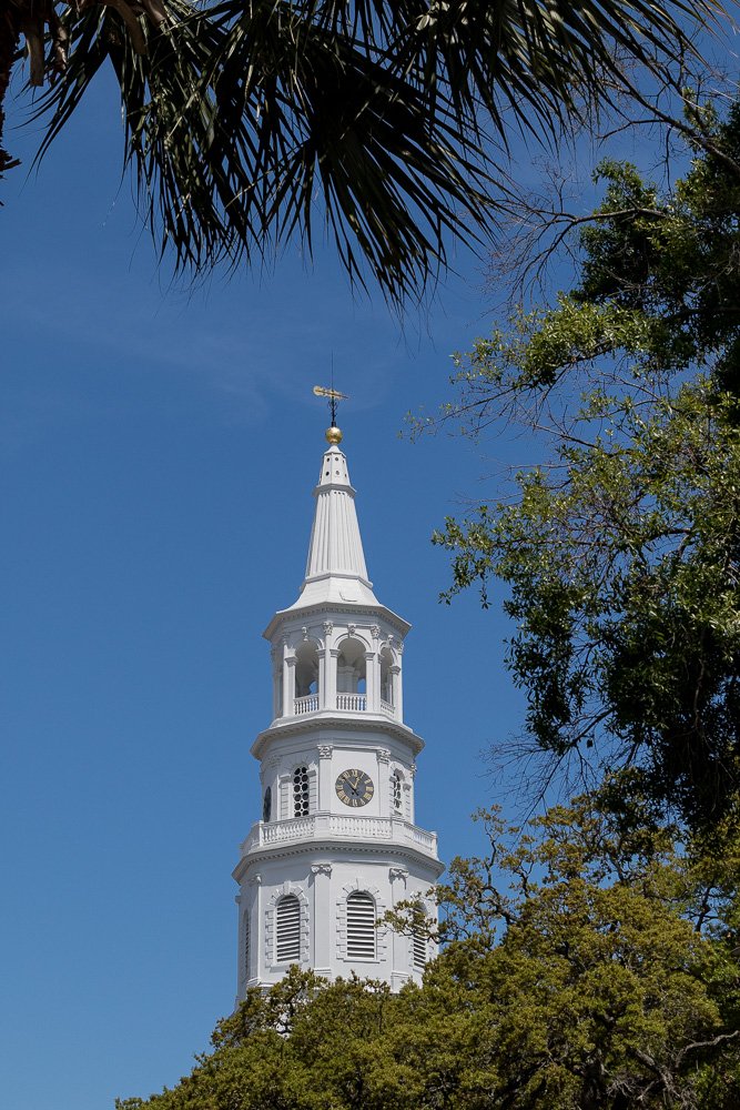 A white church steeple with a clock, surrounded by green trees and a blue sky. St. Michael's Episcopal Church. Photography Classes in Charleston SC. Camera Classes in Charleston SC
