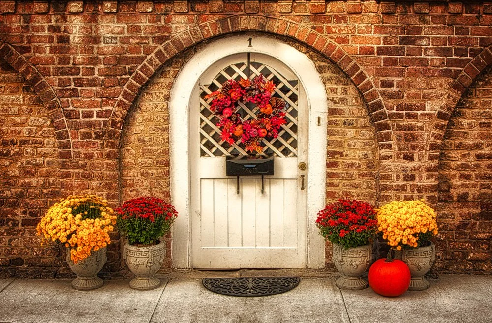 A white door with a heart-shaped flower wreath hangs on a brick wall. Potted yellow and red chrysanthemums are on either side of the door, with an orange pumpkin on the right. Photography Classes in Charleston SC. Camera Classes in Charleston SC