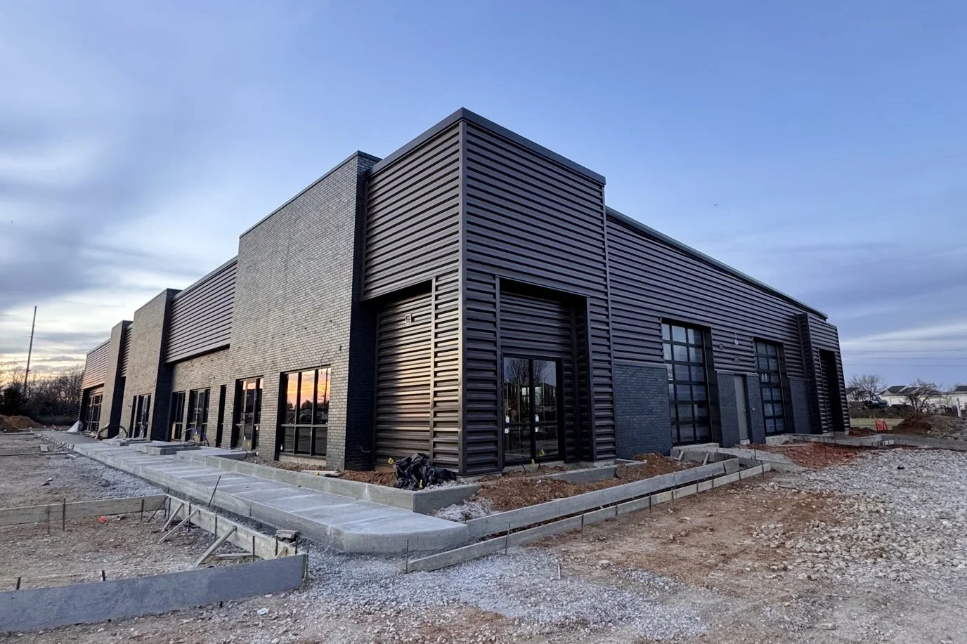 Newly constructed modern commercial building with black metal siding and large windows, under construction with a gravel and dirt surrounding area at sunset.