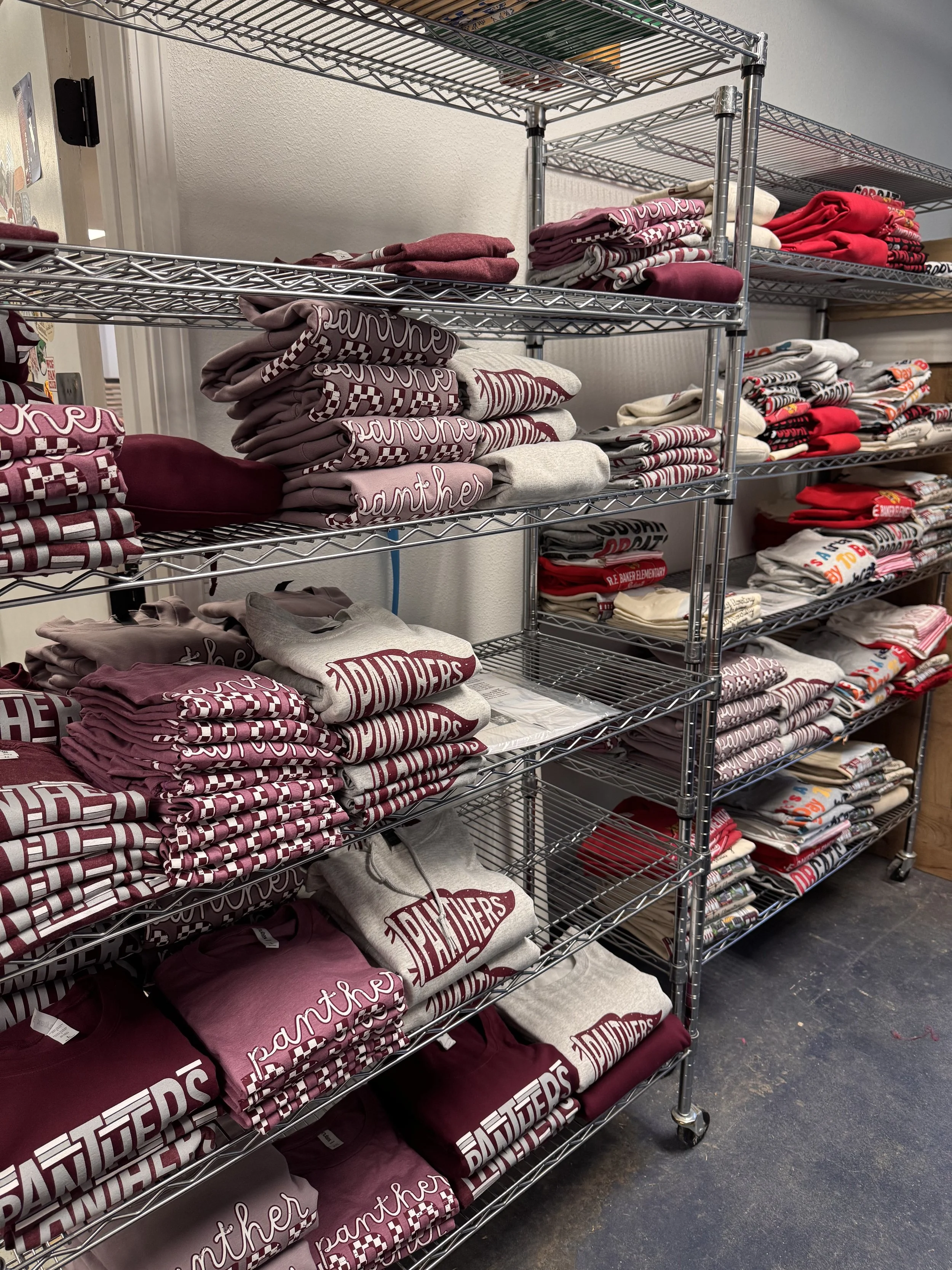 Metal shelving units filled with folded maroon, white, and gray t-shirts and sweatshirts with various designs, on wheels, situated on a gray floor.