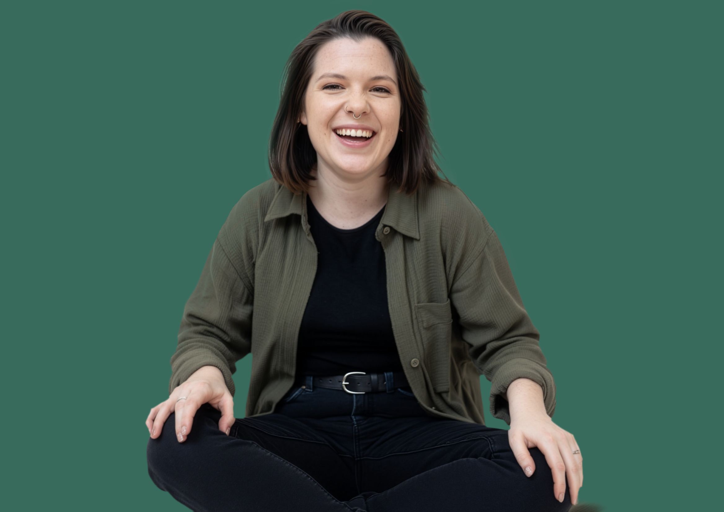 A young woman with shoulder-length brown hair, smiling, sitting cross-legged against a plain dark green background, wearing a black t-shirt and an open olive green shirt.