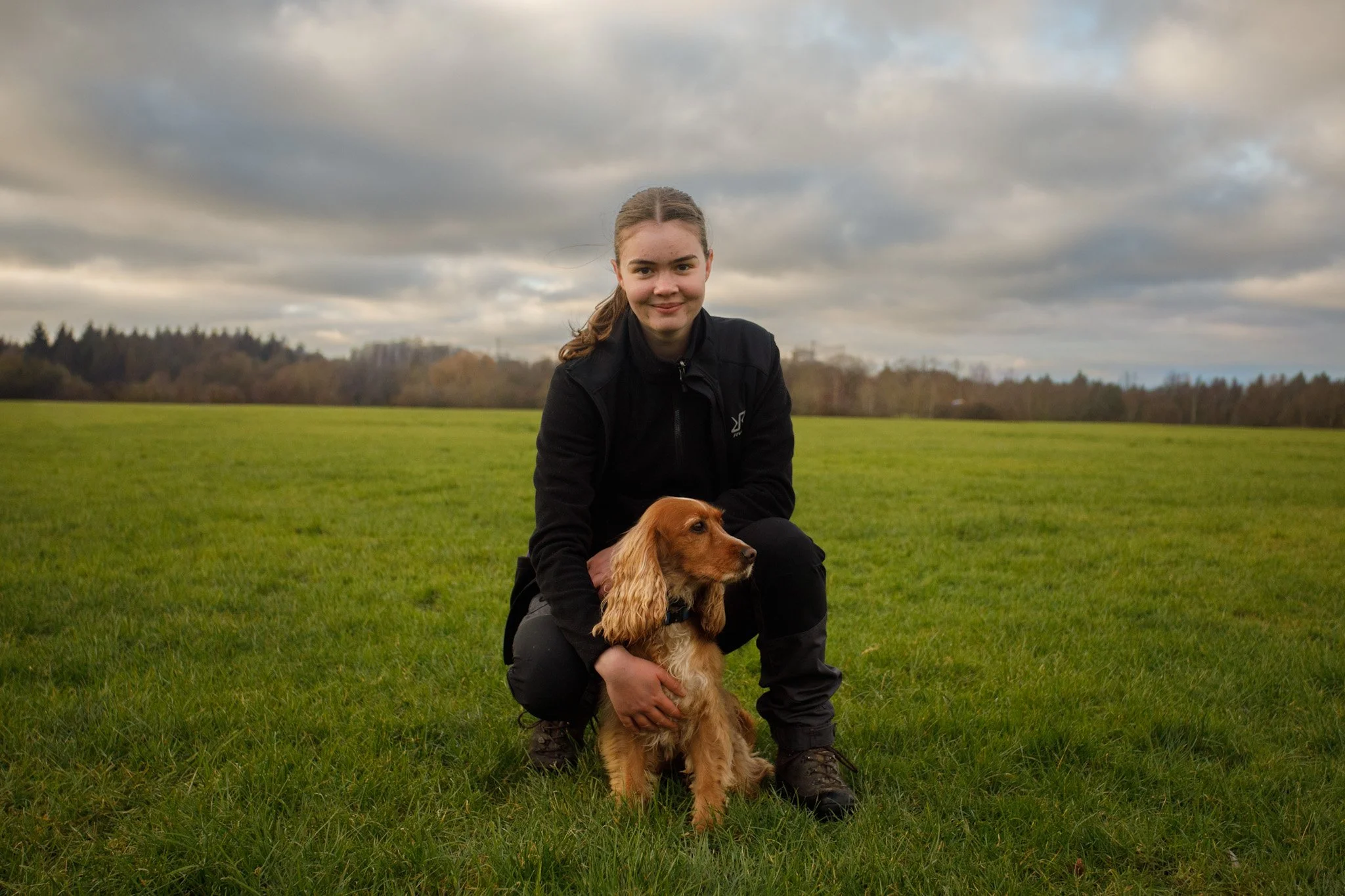 A young woman with brown hair in a ponytail, wearing a black jacket, crouches on green grass with a golden Cocker Spaniel in a scenic outdoor field under a cloudy sky.