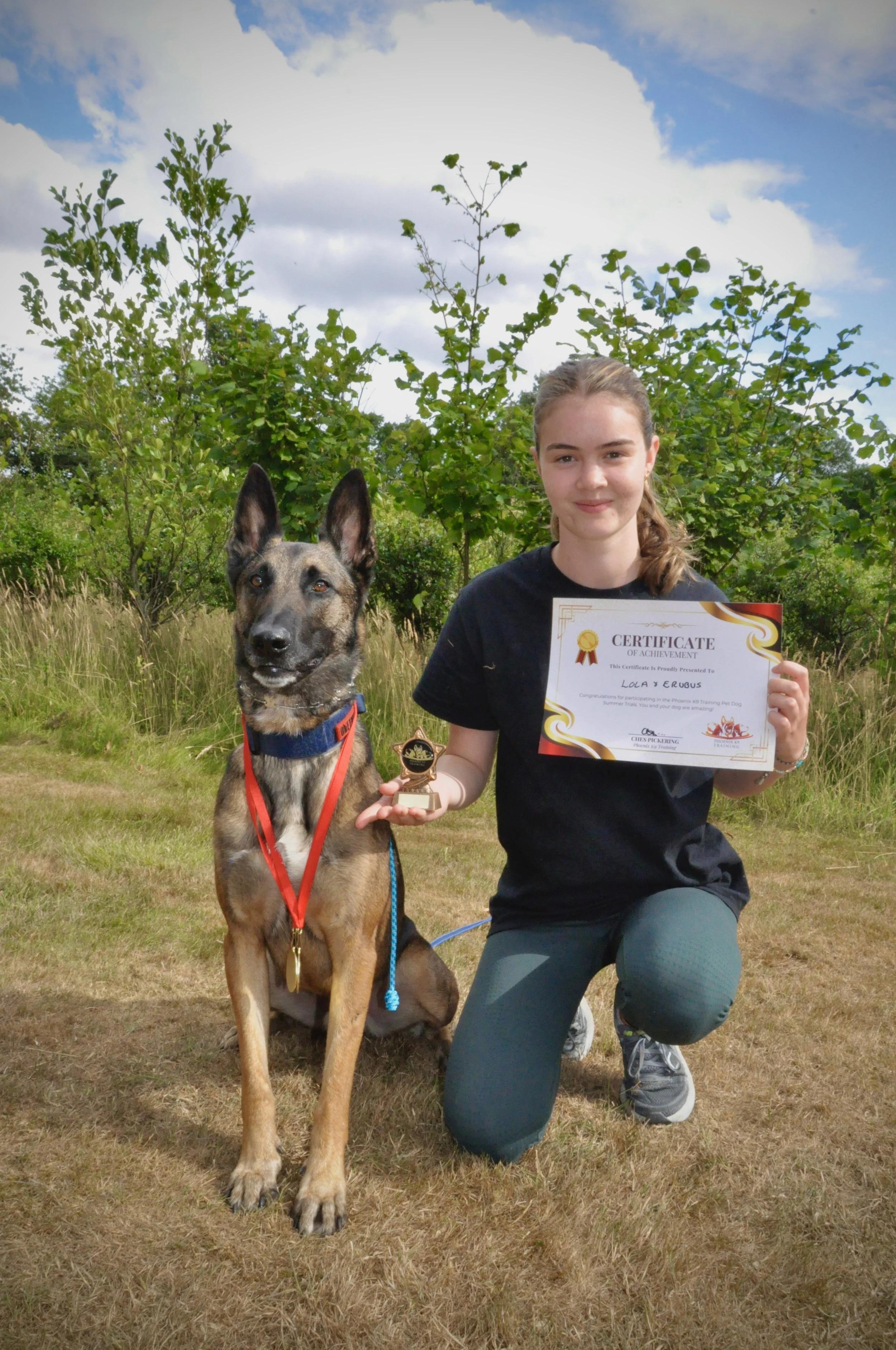 A young woman kneeling beside a Malinois dog outdoors, holding a certificate and a small trophy, both celebrating an achievement in training or competition. The scene is set in a grassy area with trees and a partly cloudy sky in the background.