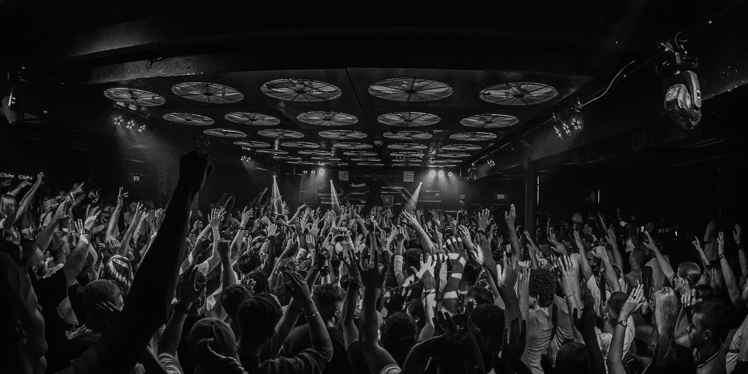 Crowd of people at a concert or nightclub dancing and raising their hands with blue stage lighting.