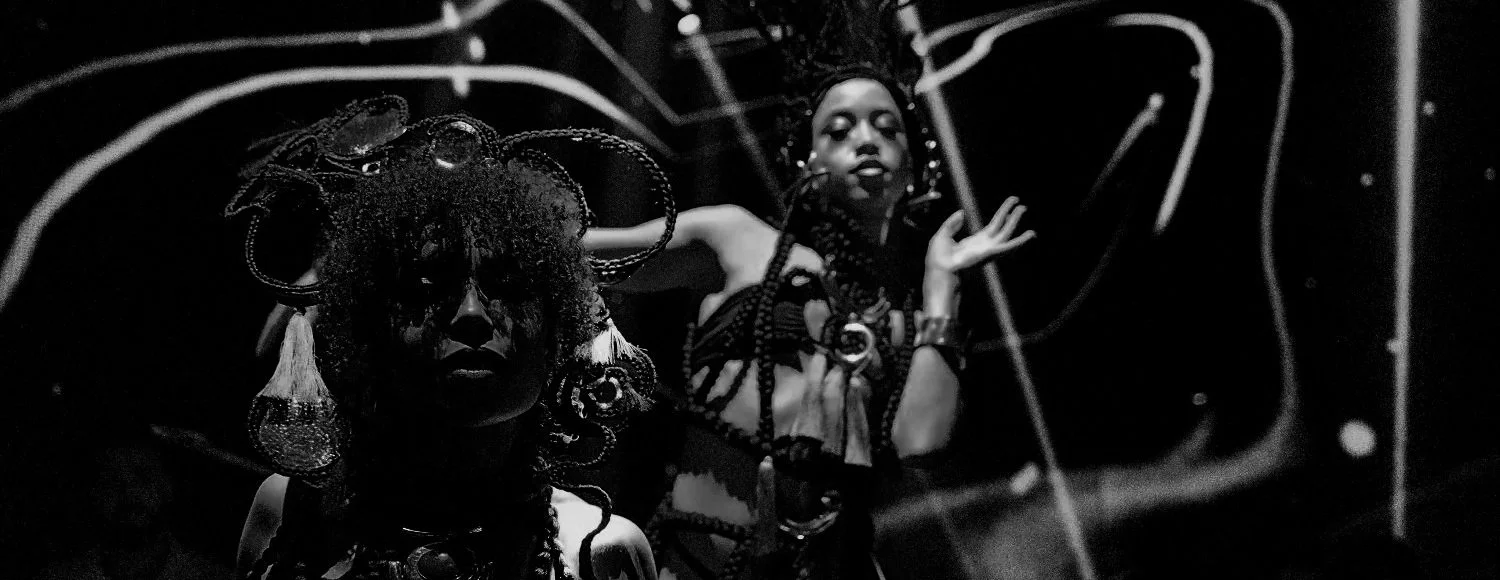 Two women dressed in elaborate, traditional African-style costumes with intricate beadwork and jewelry, posing against a dark background with abstract light patterns.