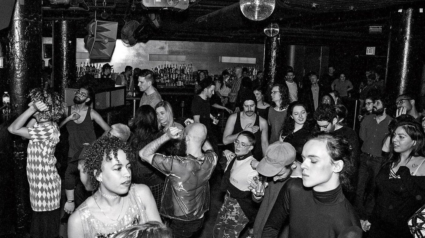 Crowd of people dancing and socializing in a nightclub or bar with a bar area in the background.