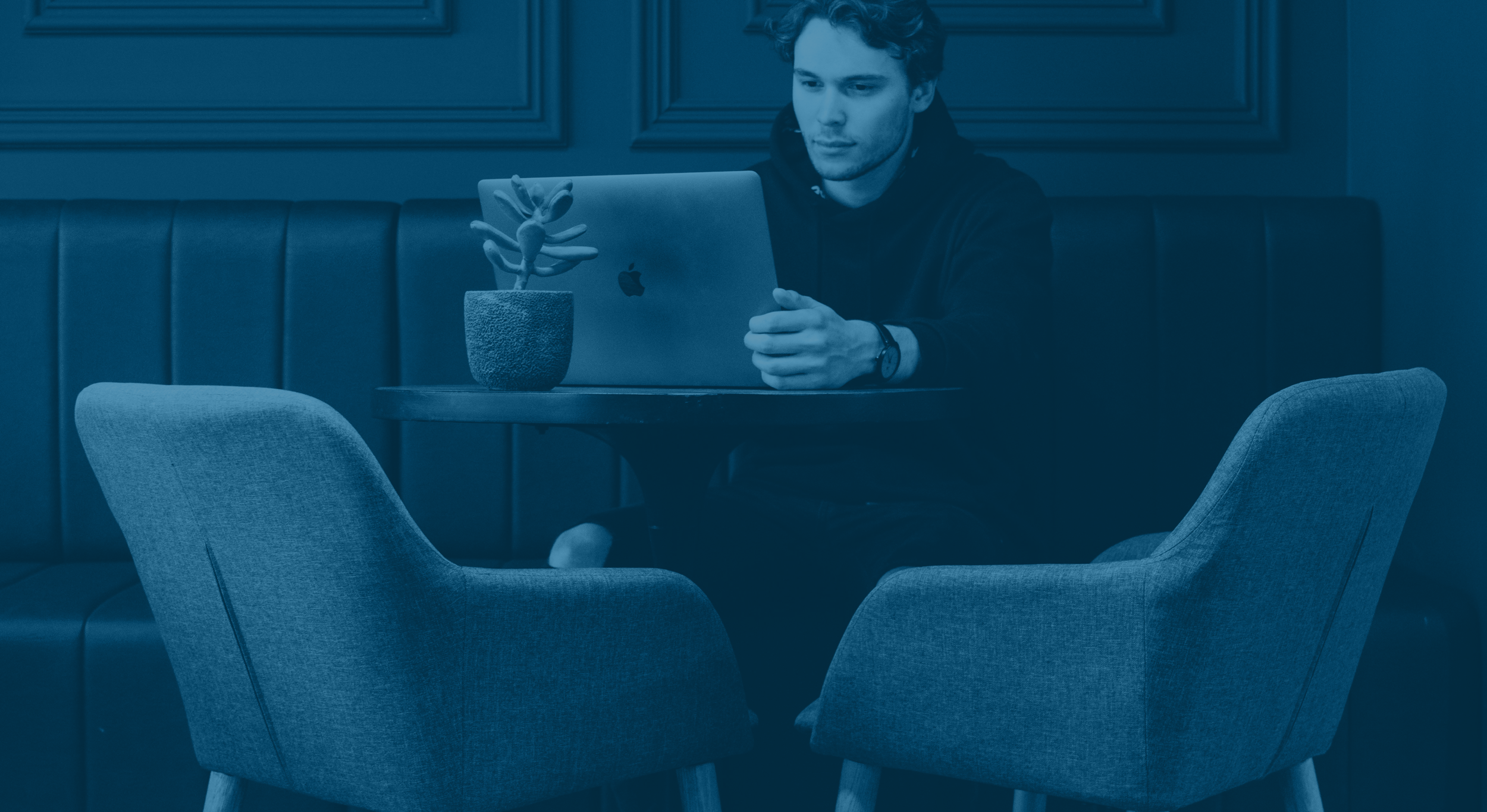 A young man with curly hair sitting at a dark wooden table in a cafe or restaurant, looking at a laptop with an Apple logo, with a potted succulent plant on the table, surrounded by upholstered chairs and wood-paneled walls.