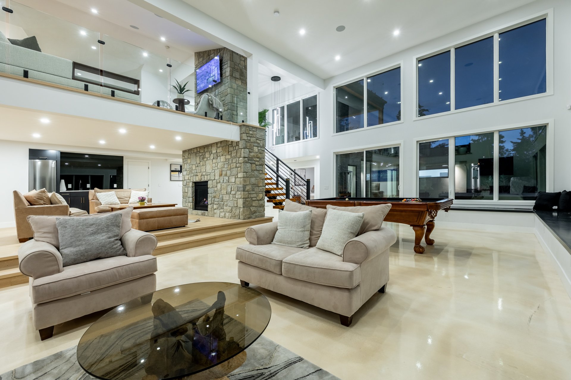 Spacious modern living room with large windows, beige sofas, a stone fireplace, a pool table, and a staircase leading to a second level.