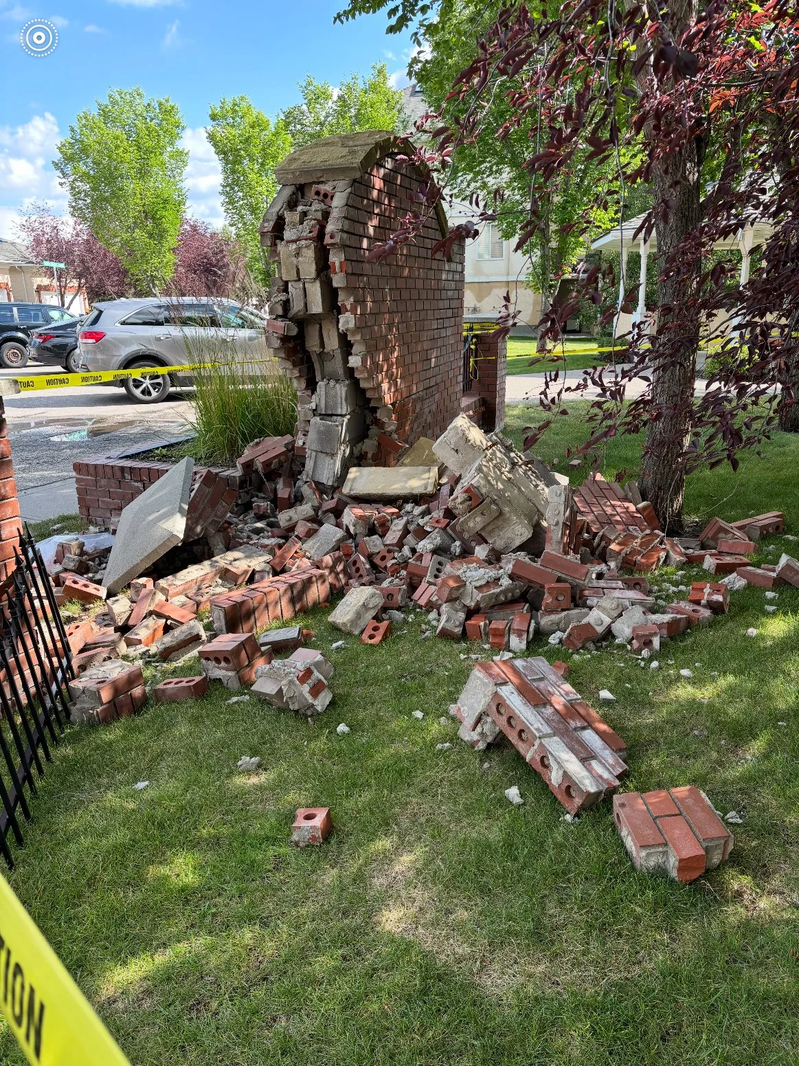 A brick wall on a house has collapsed onto a lawn, with broken bricks and debris scattered around, and yellow caution tape cordoning off the area.