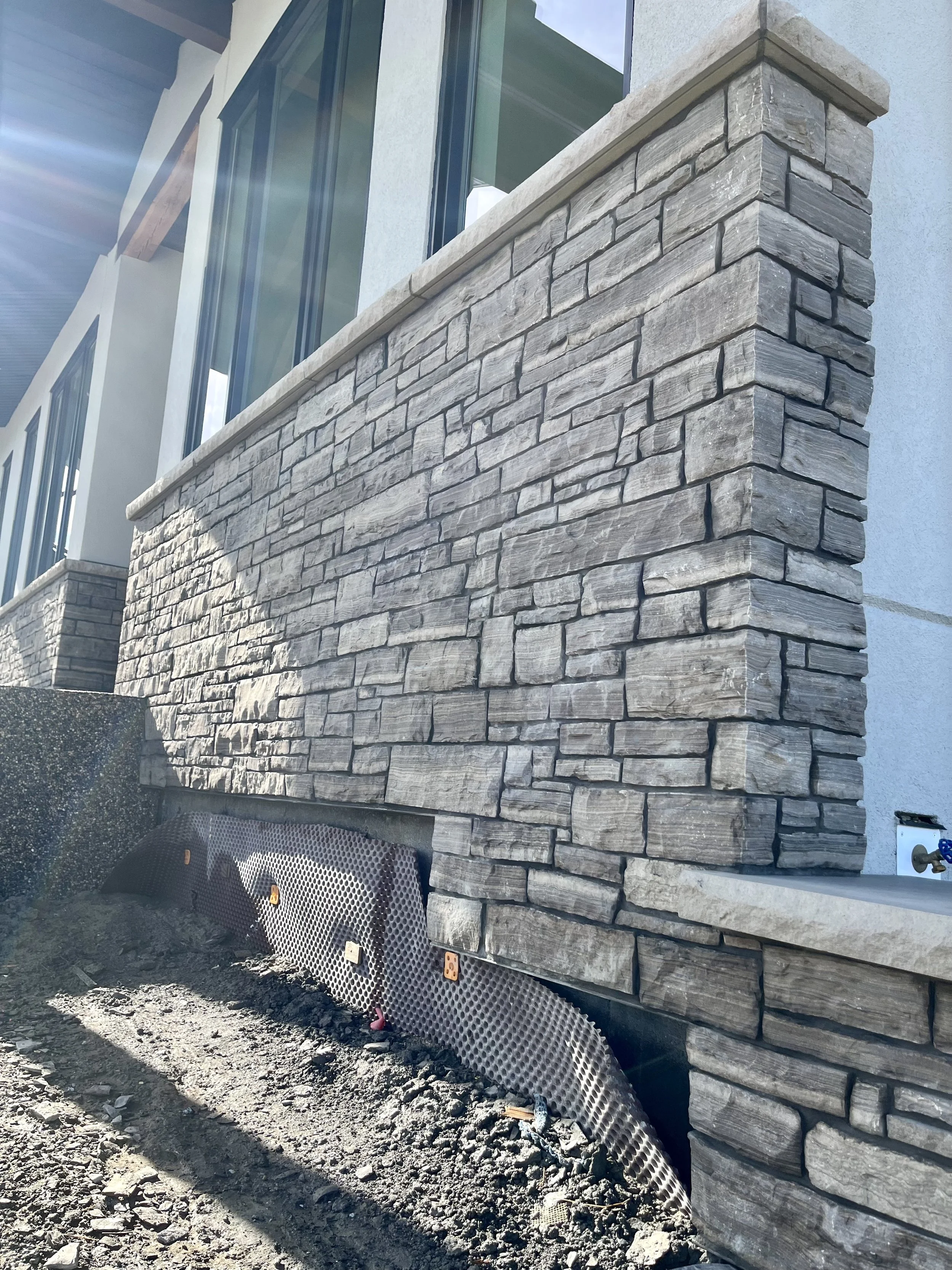 Stone veneer siding on a building exterior with a drainage system at the base, seen under bright sunlight.