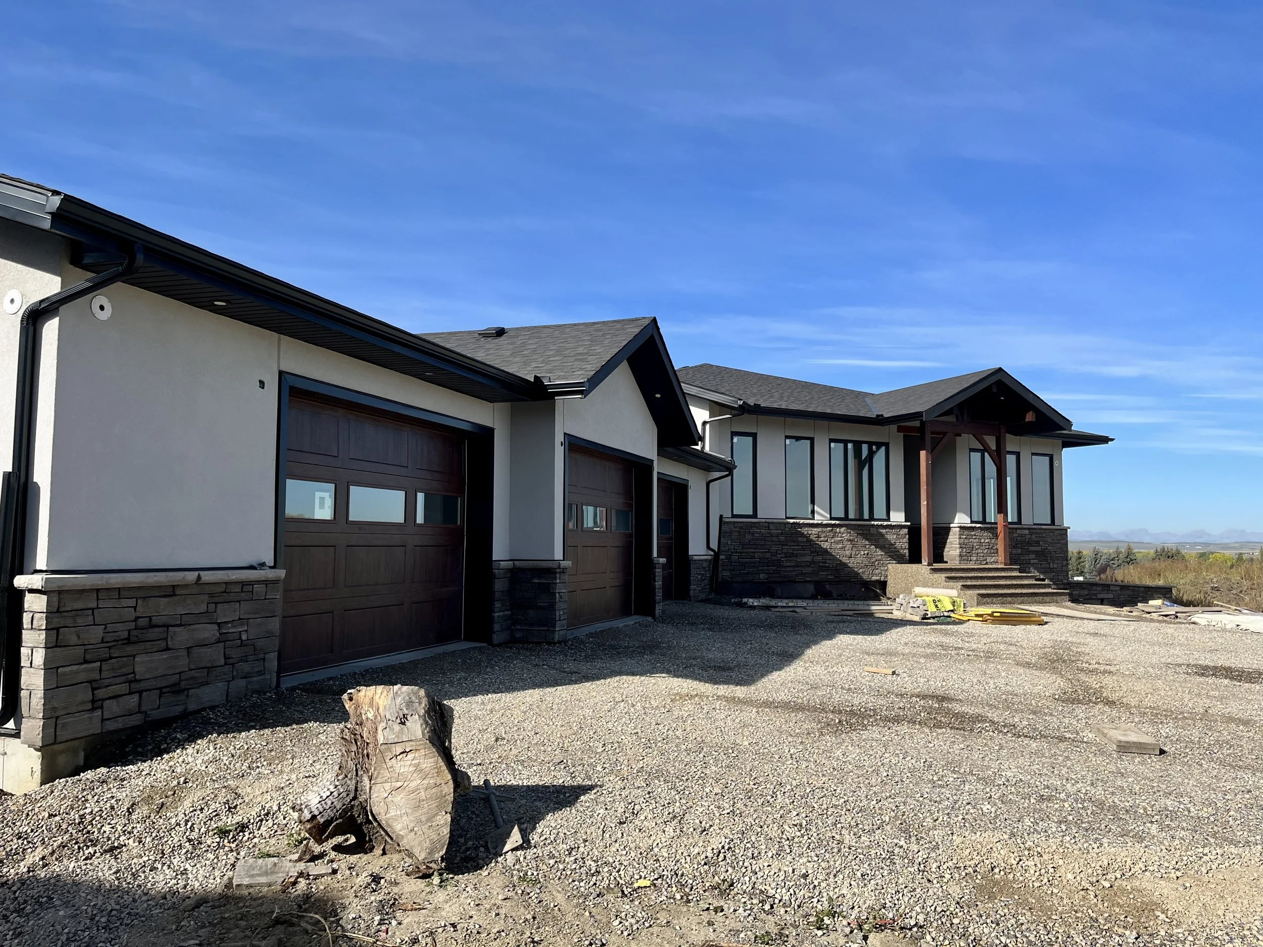 Newly constructed modern house with a two-car garage, rock facade, and gravel driveway under a blue sky.