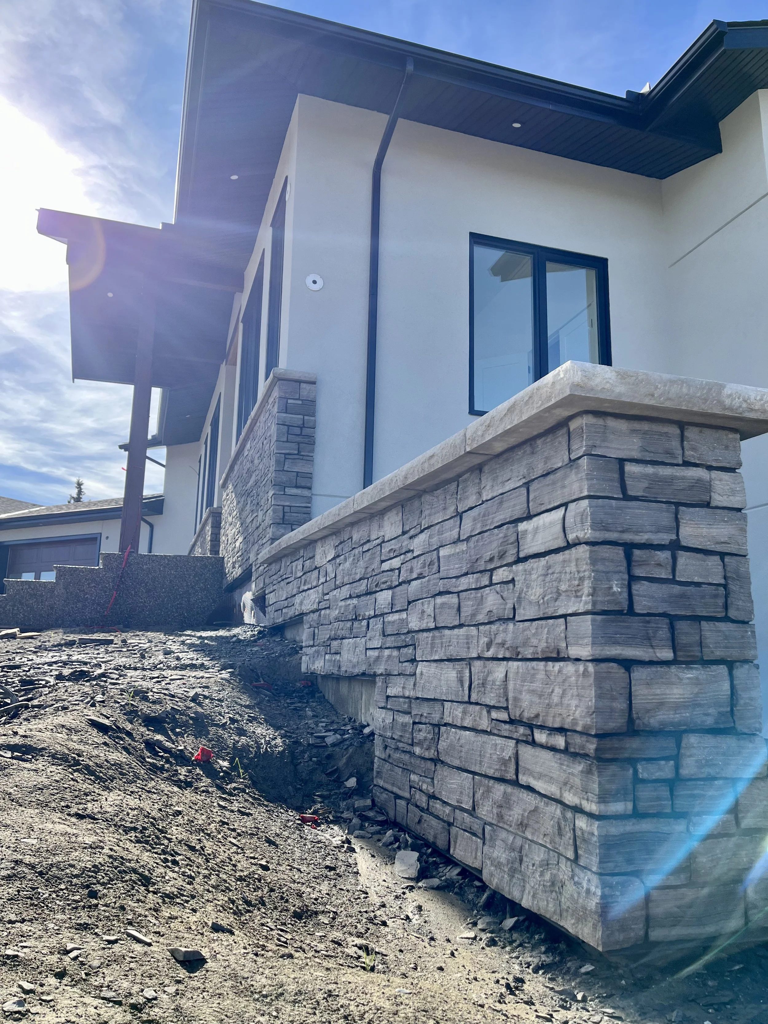 Modern house exterior with stone facade and large windows, bright sunlight in the background.