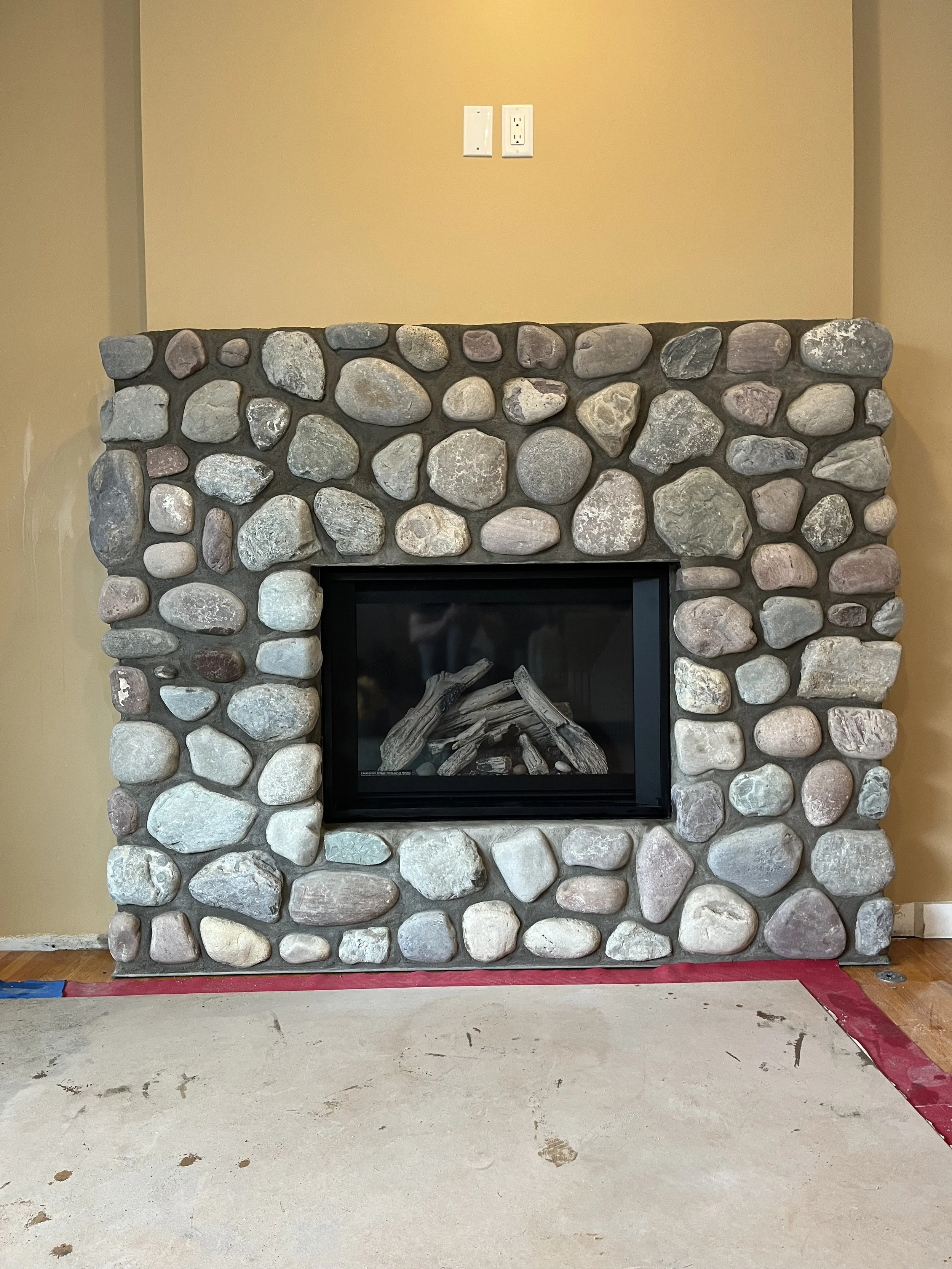 Stone fireplace with logs inside, set against a beige wall and electrical outlets above.