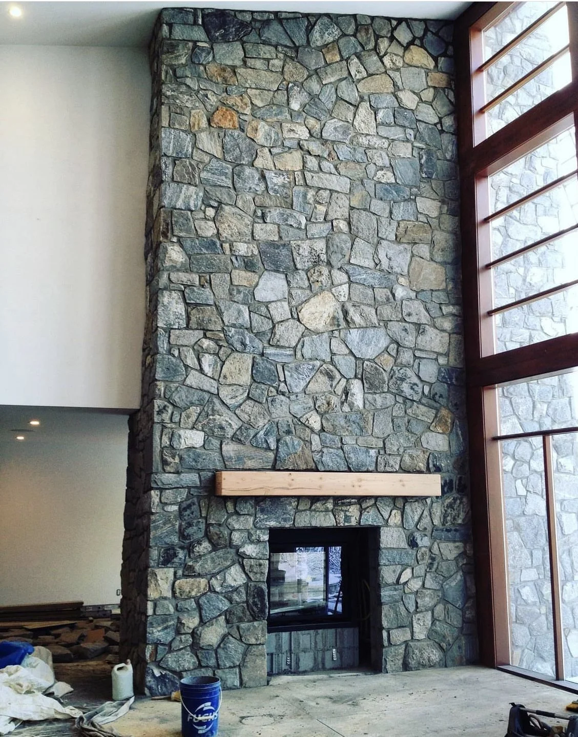 Stone fireplace with wooden mantle under construction in a modern living room with large windows.