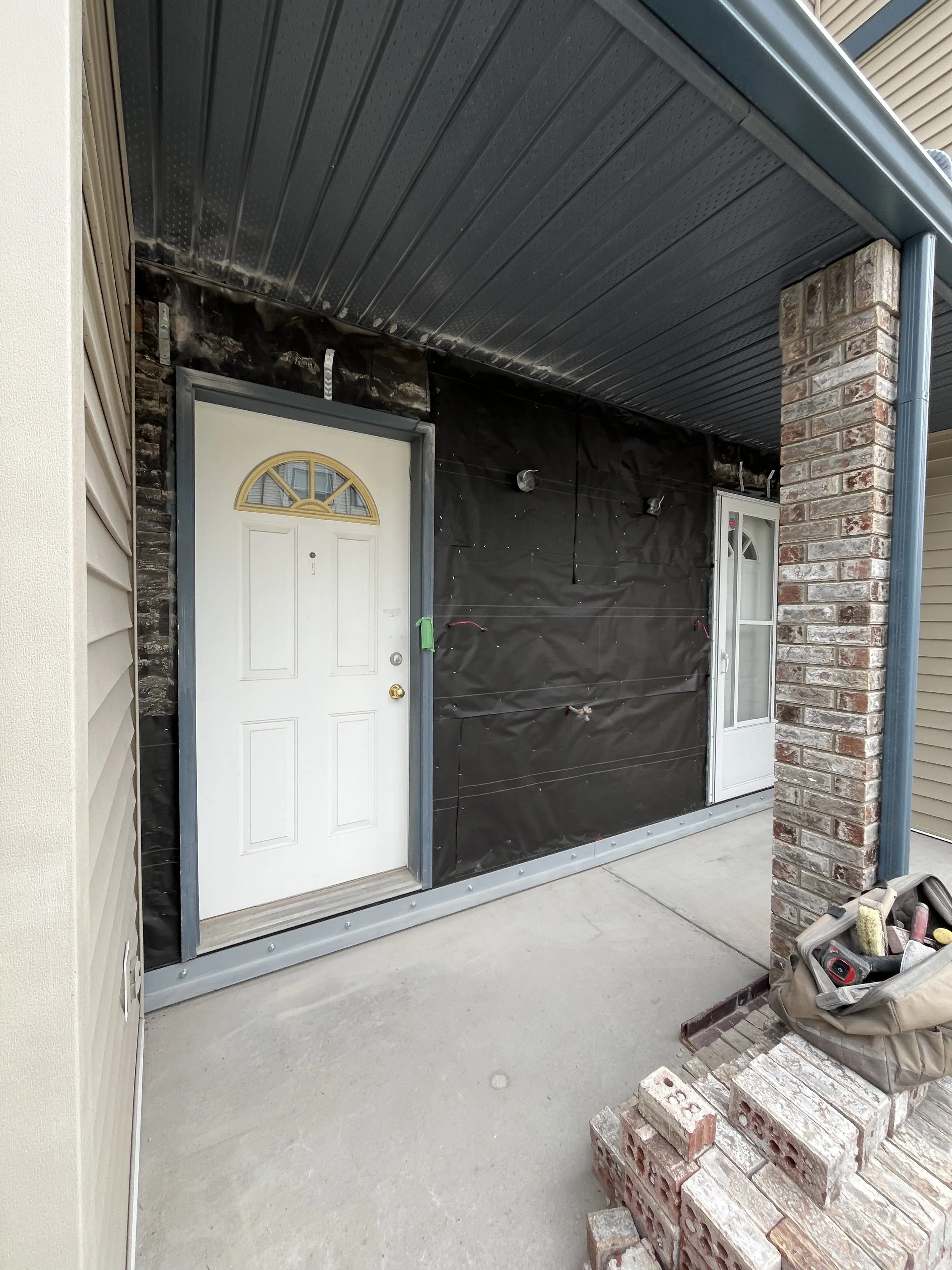 House exterior under renovation with new white doors, black protective wrap, bricks stacked, and construction tools on porch.
