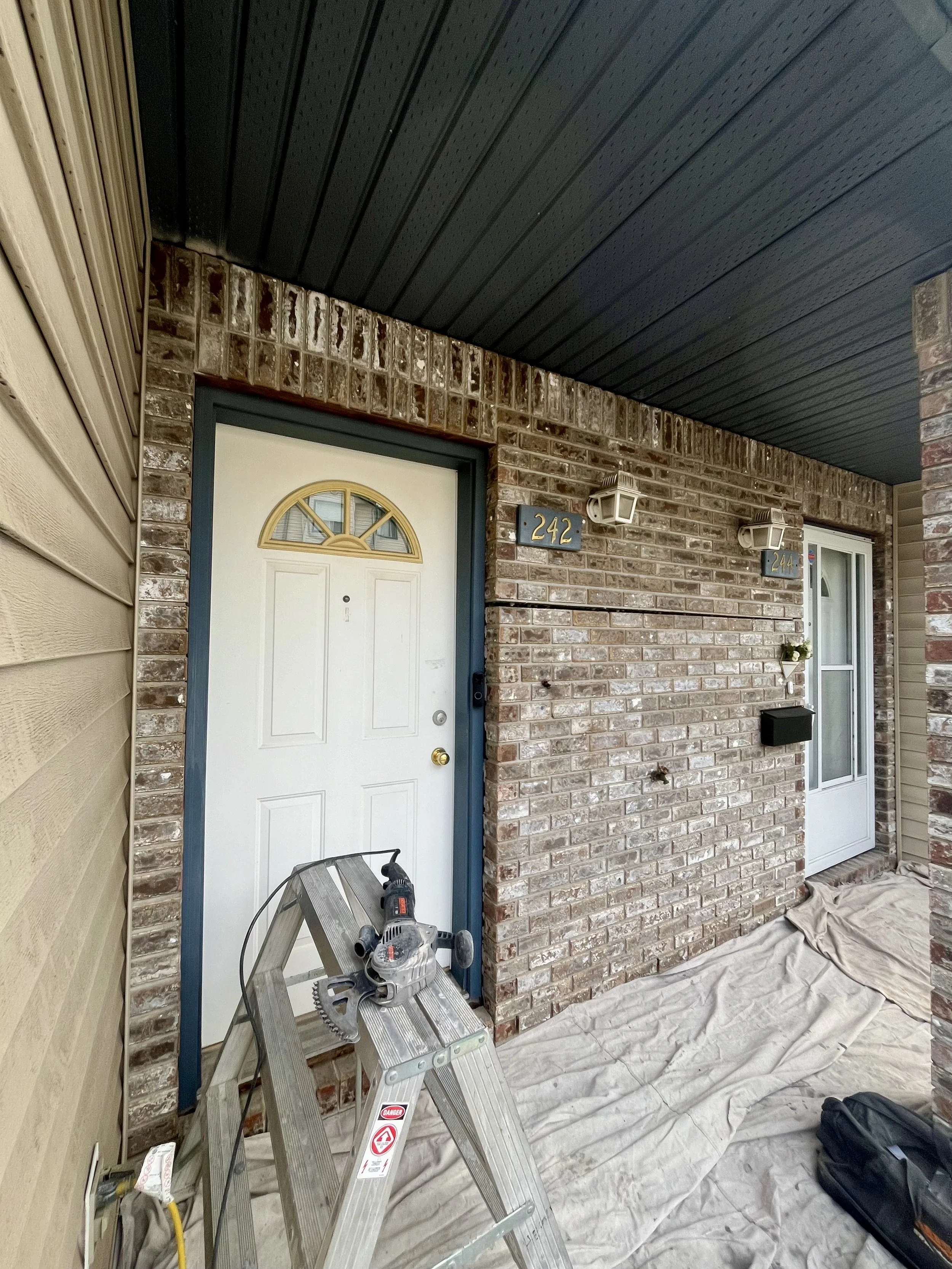 Front porch with a white door, brick walls, house number 242, a metal ladder, power tool, and drop cloths on the floor.