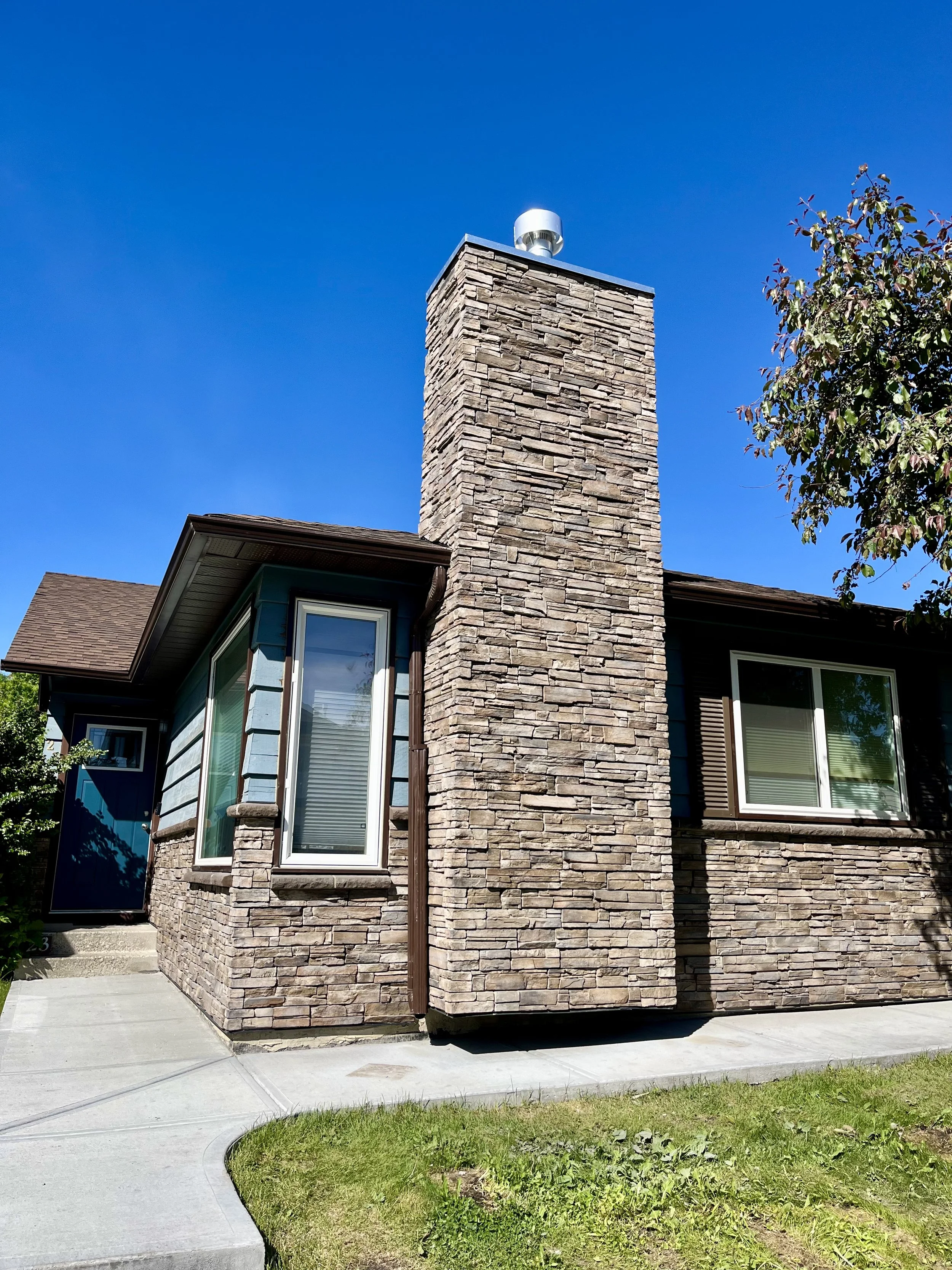 Residential house with stone exterior, large chimney, and blue siding against a clear blue sky