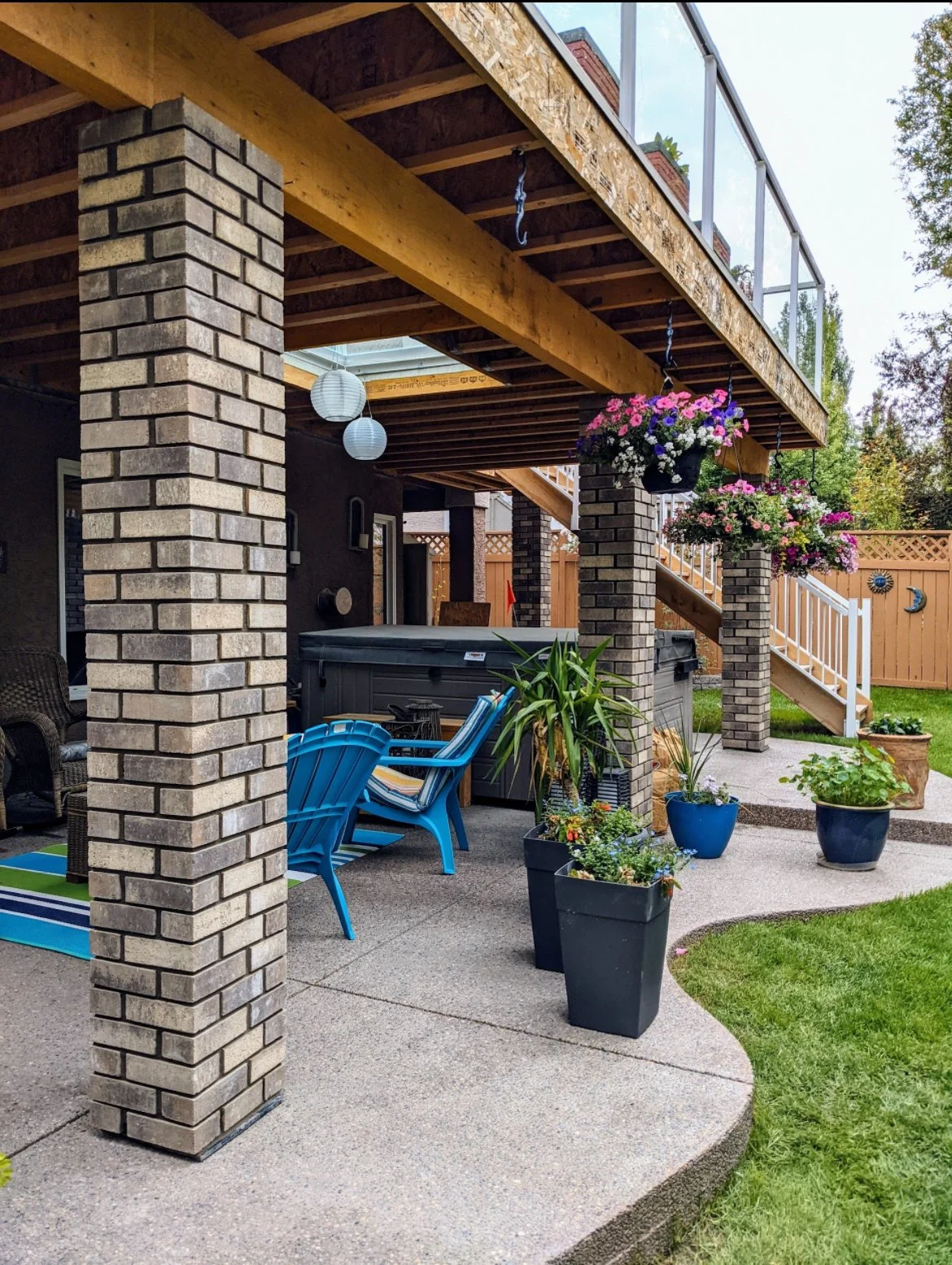Outdoor patio with brick pillars, wooden beams, blue chairs, potted plants, hanging flower baskets, and a hot tub under a deck with glass railings.