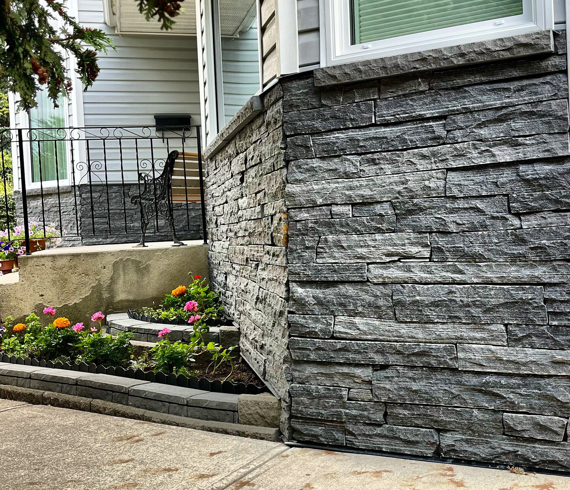 House exterior with stone veneer siding, a small garden with colorful flowers, a metal railing, and a window.