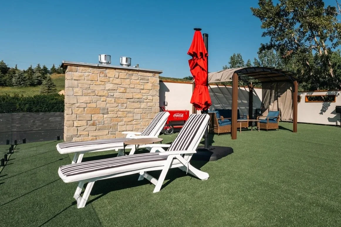 Backyard patio with two lounge chairs, a red umbrella, a stone chimney, a shaded seating area with wicker furniture, and a green hedge in the background.