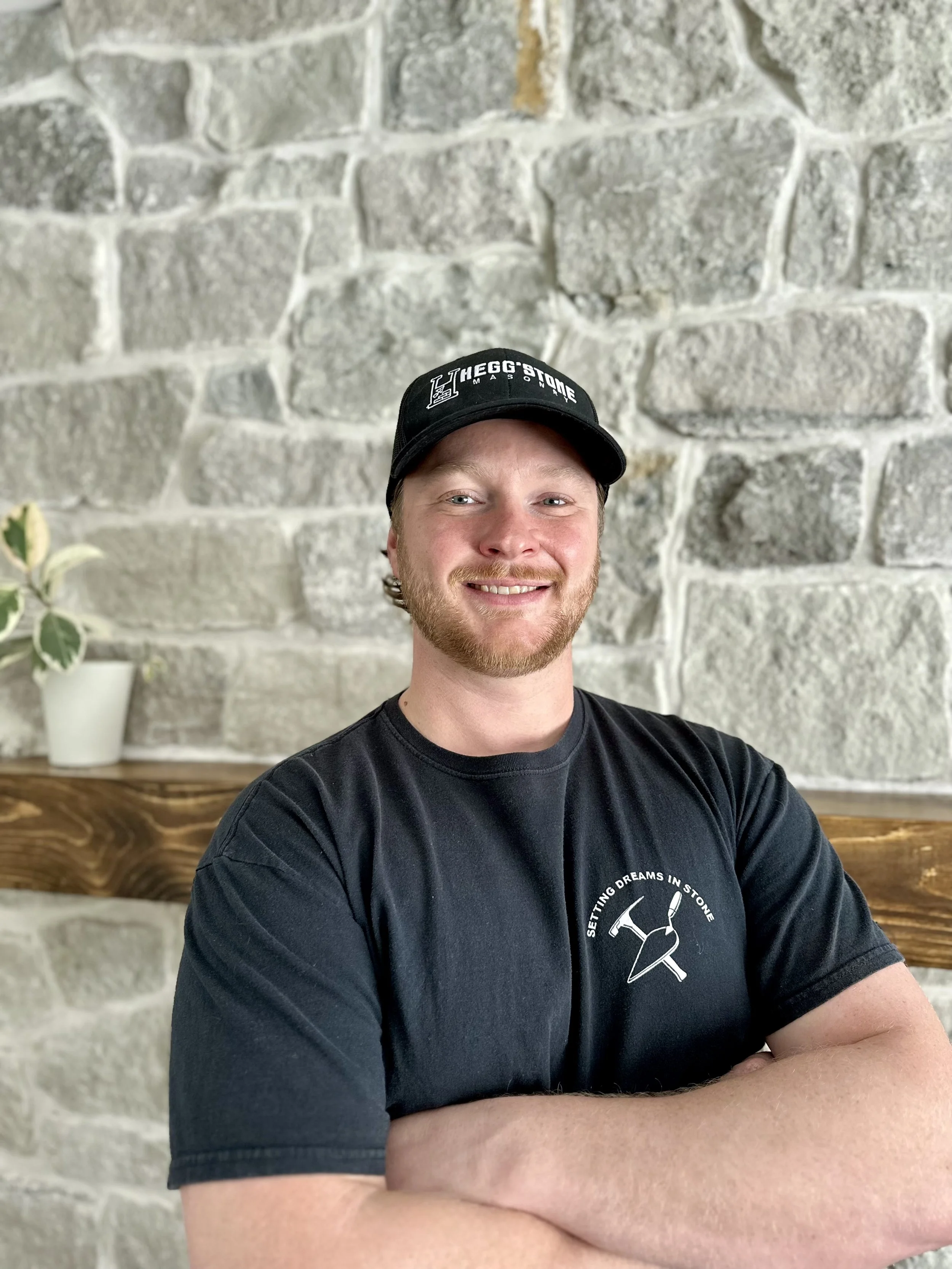 A smiling man with a beard and mustache wearing a black cap with the logo of The Gg'stone Masonry and a black T-shirt with a logo and the phrase 'Setting dreams in stone,' standing in front of a stone wall with a wooden shelf and a potted plant.