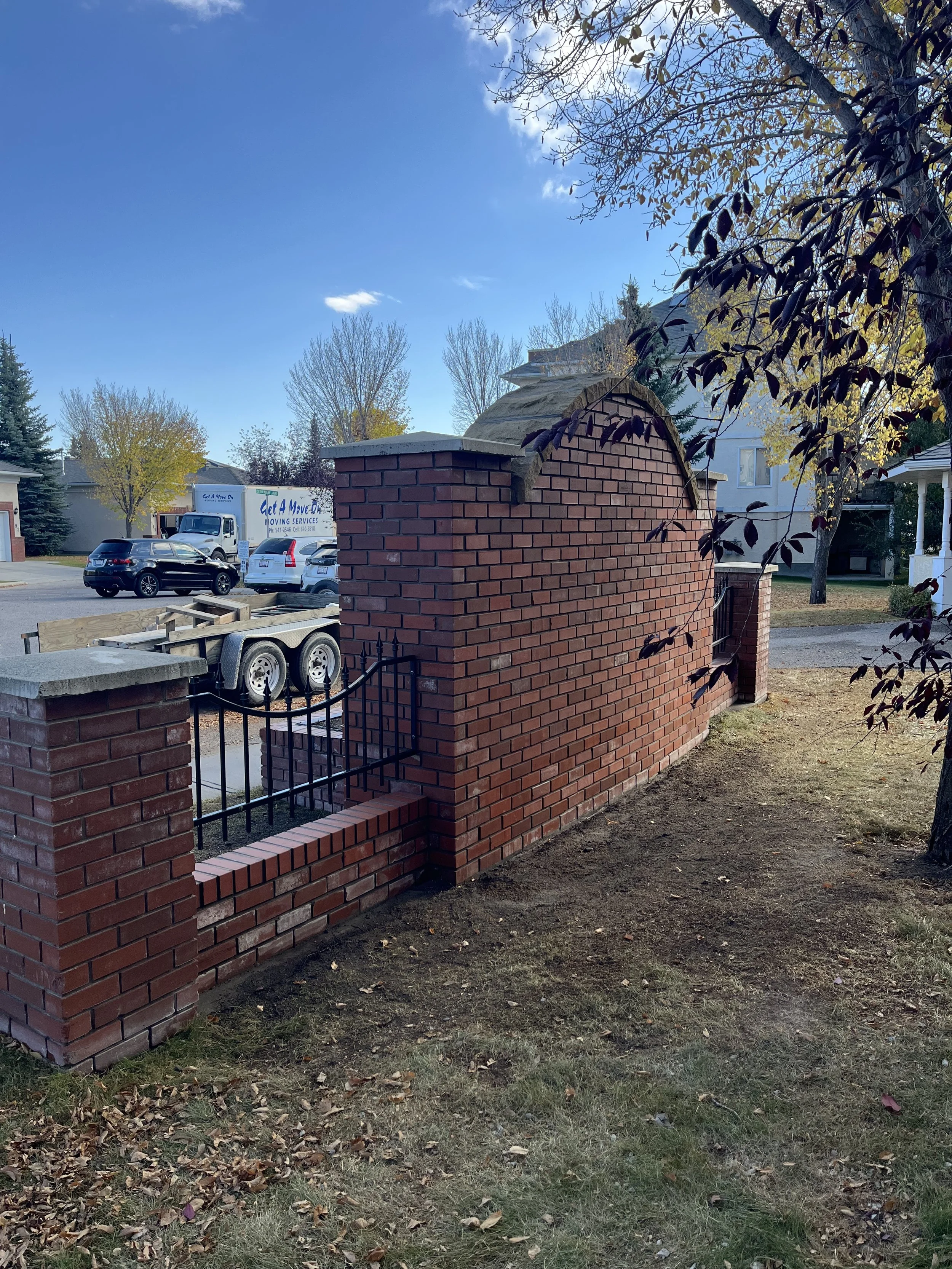 A brick wall with a black metal gate, likely part of a residential property, with a tree overhanging on the right side, and a clear blue sky with a few clouds.