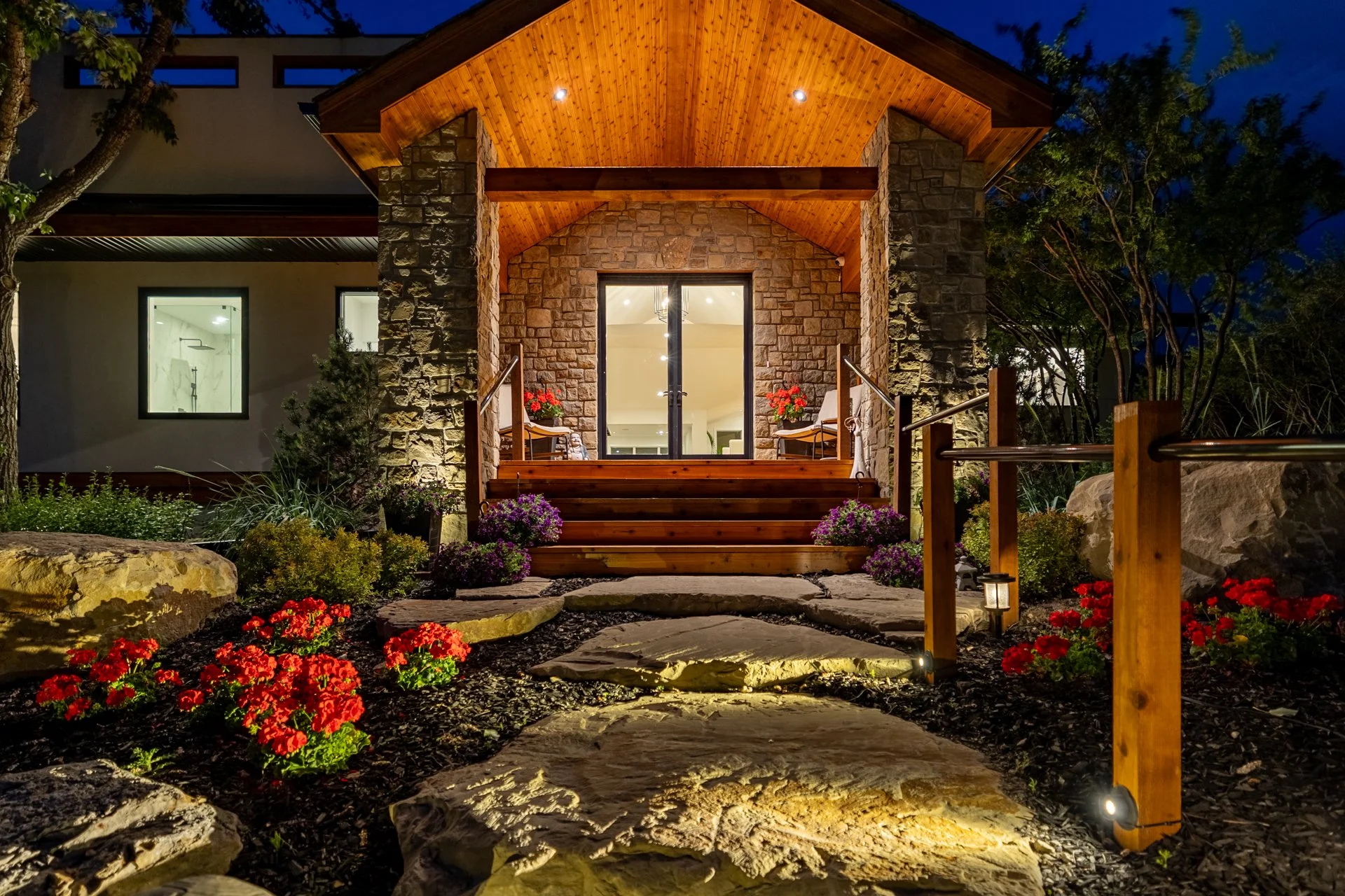 Night view of a house entrance with stone steps, wooden railing, illuminated porch, potted plants with red and purple flowers, and landscaped garden with rocks and flowers.
