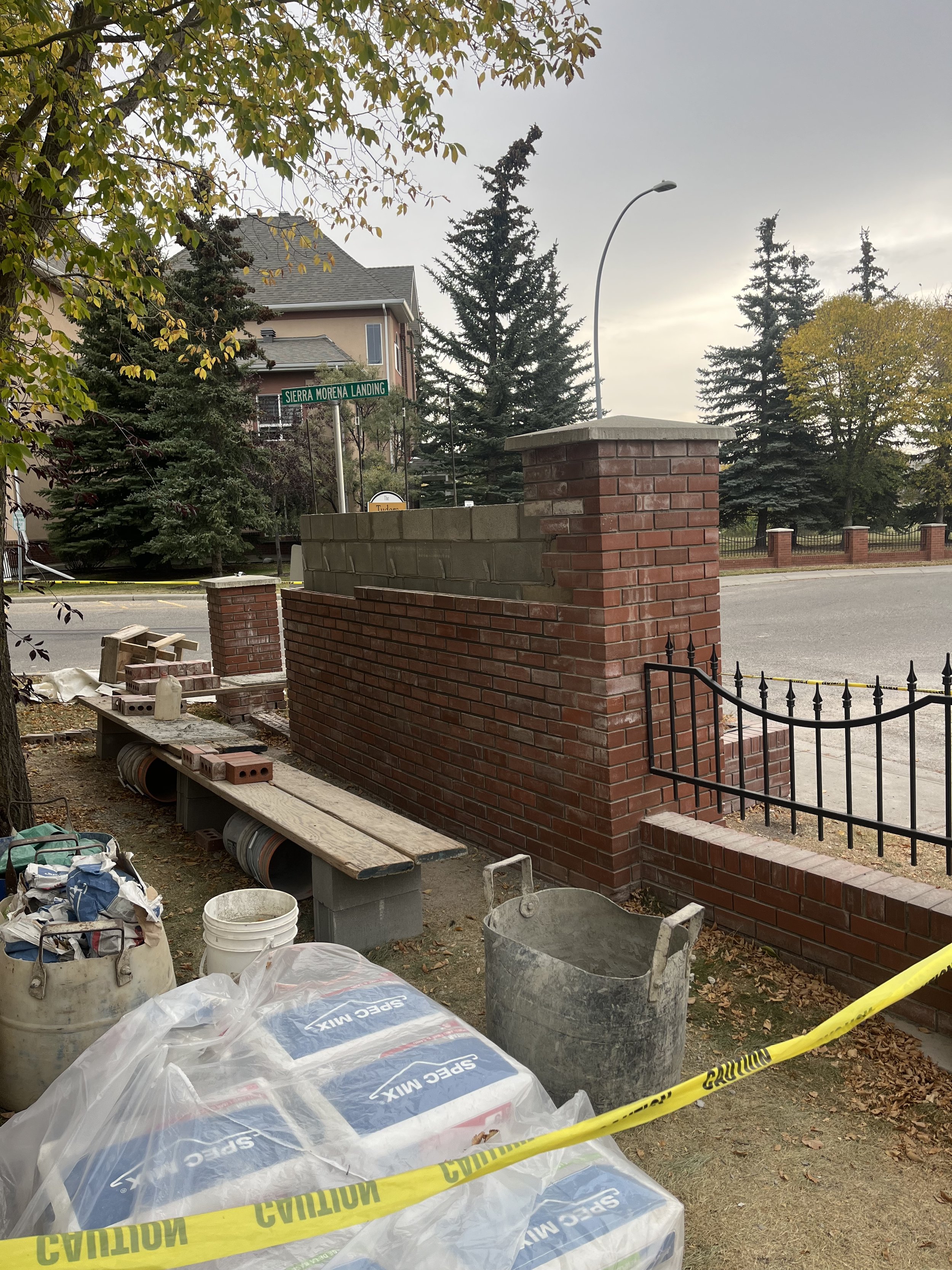A construction site with a brick wall and patio area, construction tools, and materials, yellow caution tape, trees, and a street sign that reads 'Sierra Morena Landing' in the background.