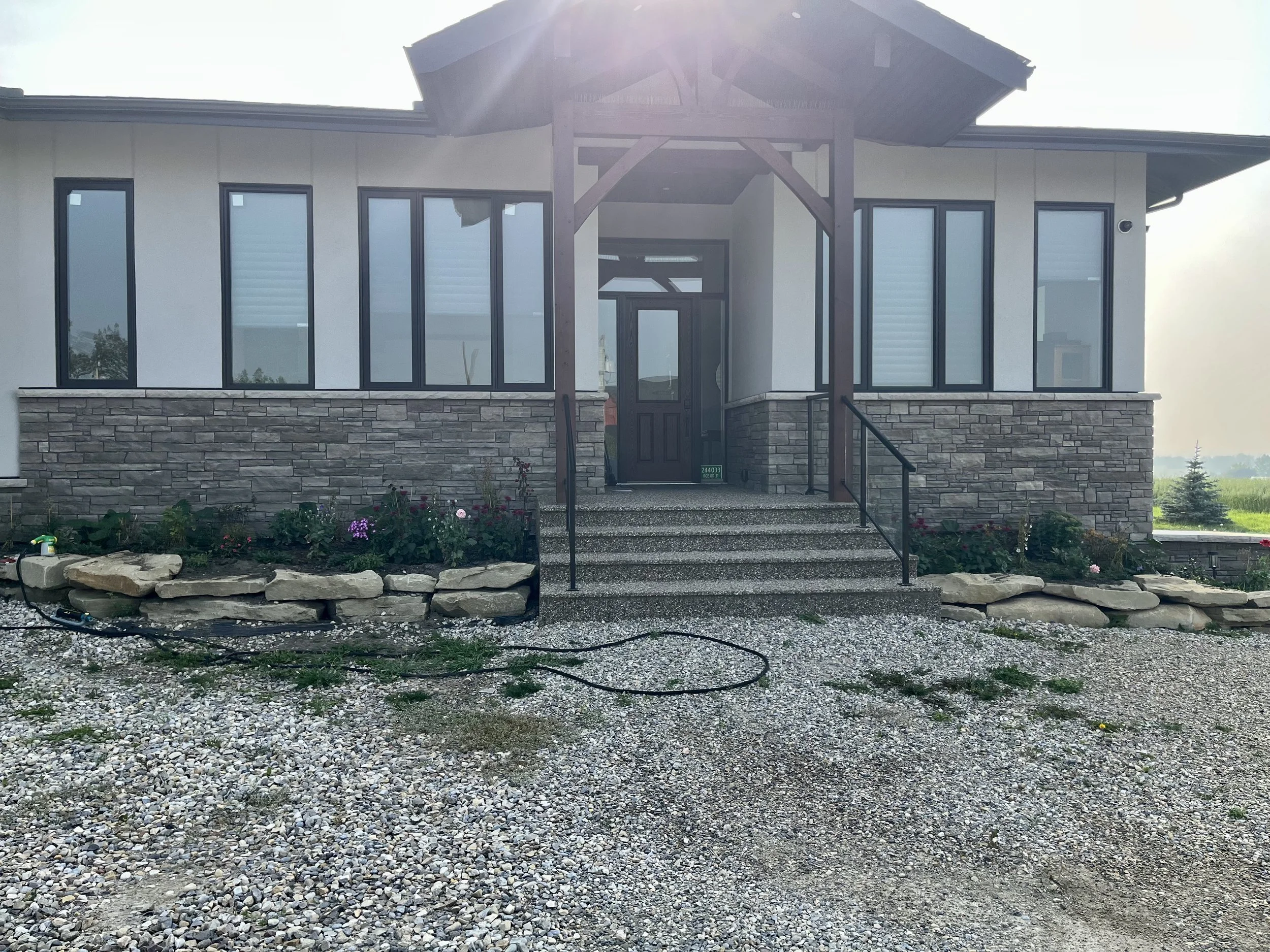 Front view of a modern house with stone and white siding exterior, large windows, a staircase leading to the front door, and landscaping with flowers and rocks.