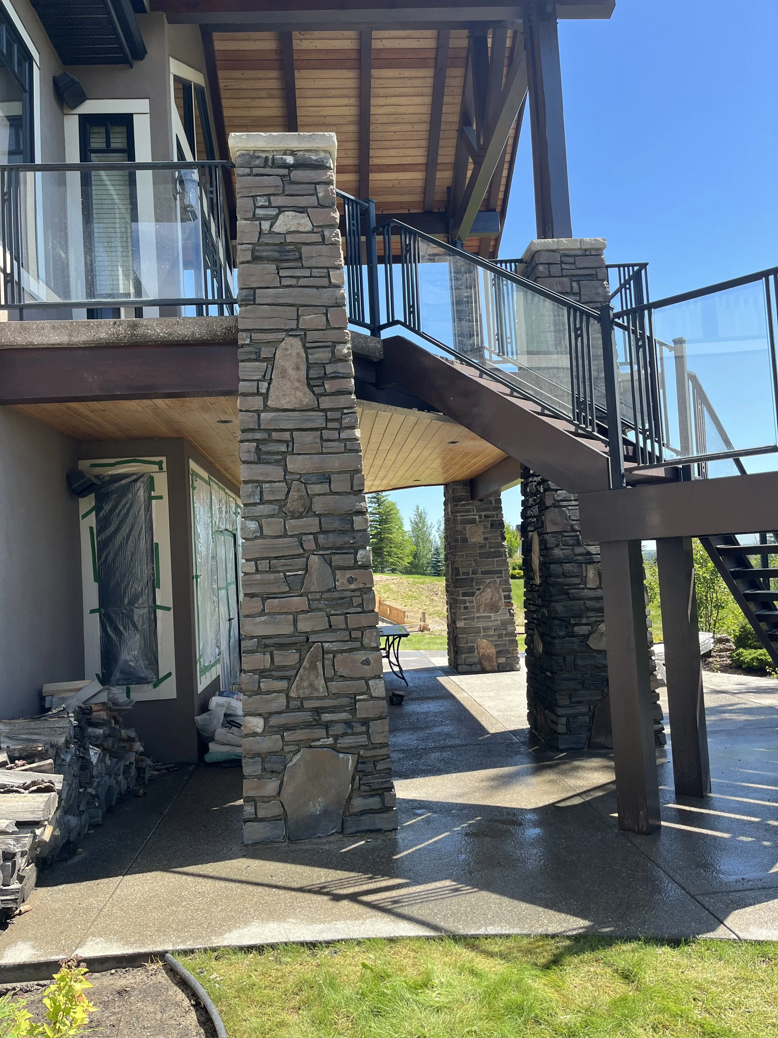 View of a house with a stone pillar and a wooden outdoor staircase leading up to a balcony with glass and metal railings. Part of the house's exterior partially covered in plastic, with construction materials nearby, and a grassy area in the backgrou