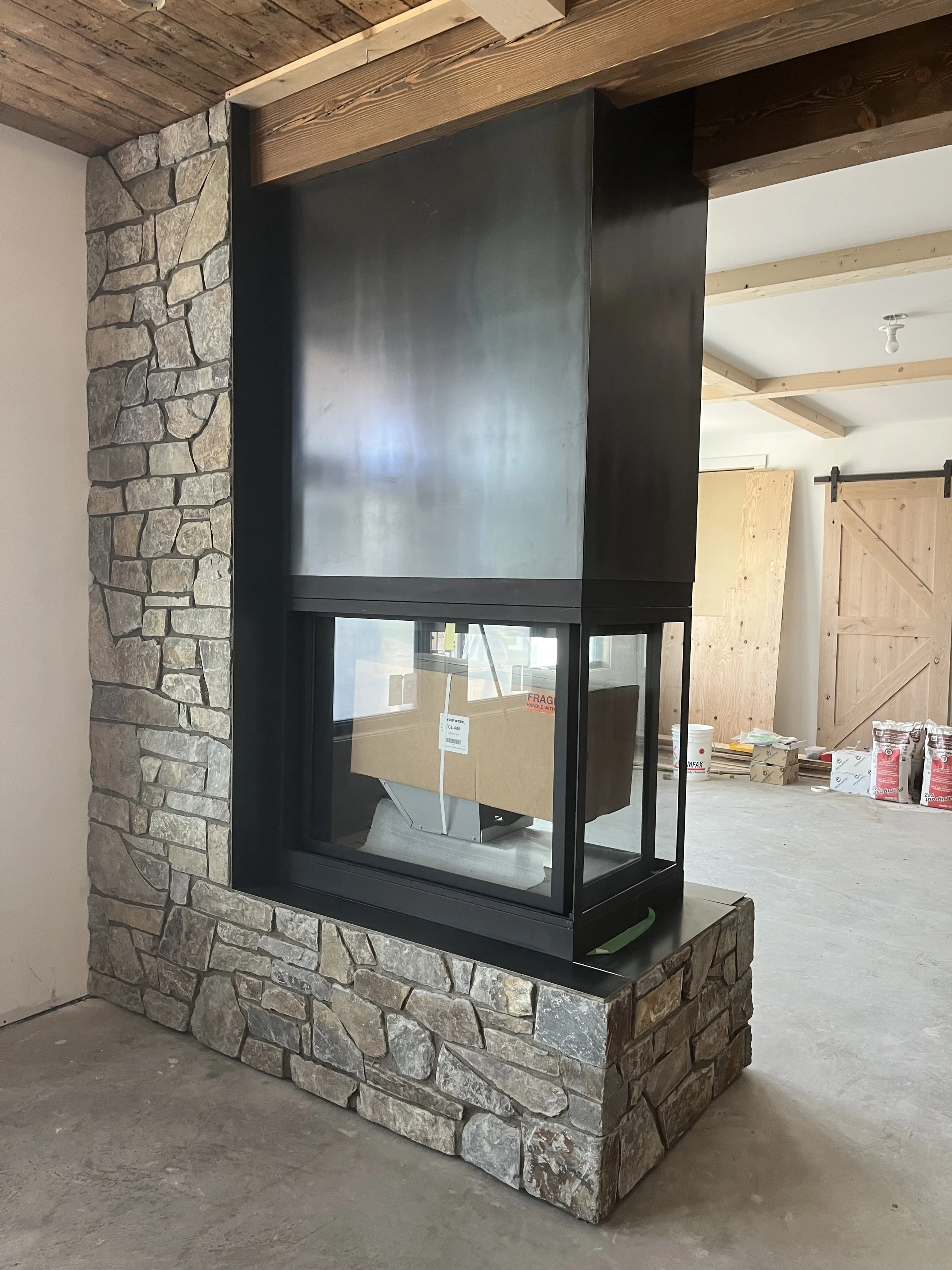 Newly installed modern fireplace with a glass enclosure, set on a stone base, in a rustic interior under construction with exposed wood beams.