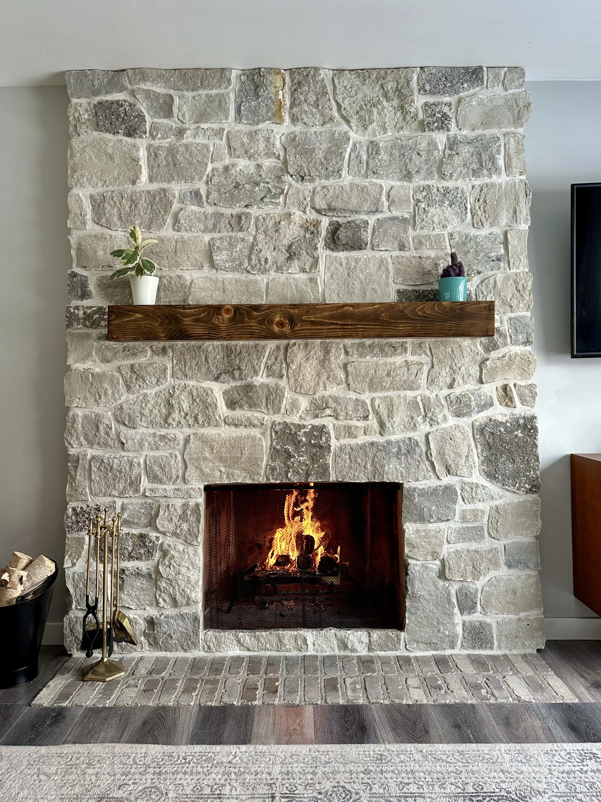 A stone fireplace with a burning fire, a wooden mantel, and two potted plants on the mantel, one with green and white leaves and the other a small purple plant in a turquoise pot. There are fireplace tools and logs beside the fireplace, and part of a gray rug and wood flooring are visible.
