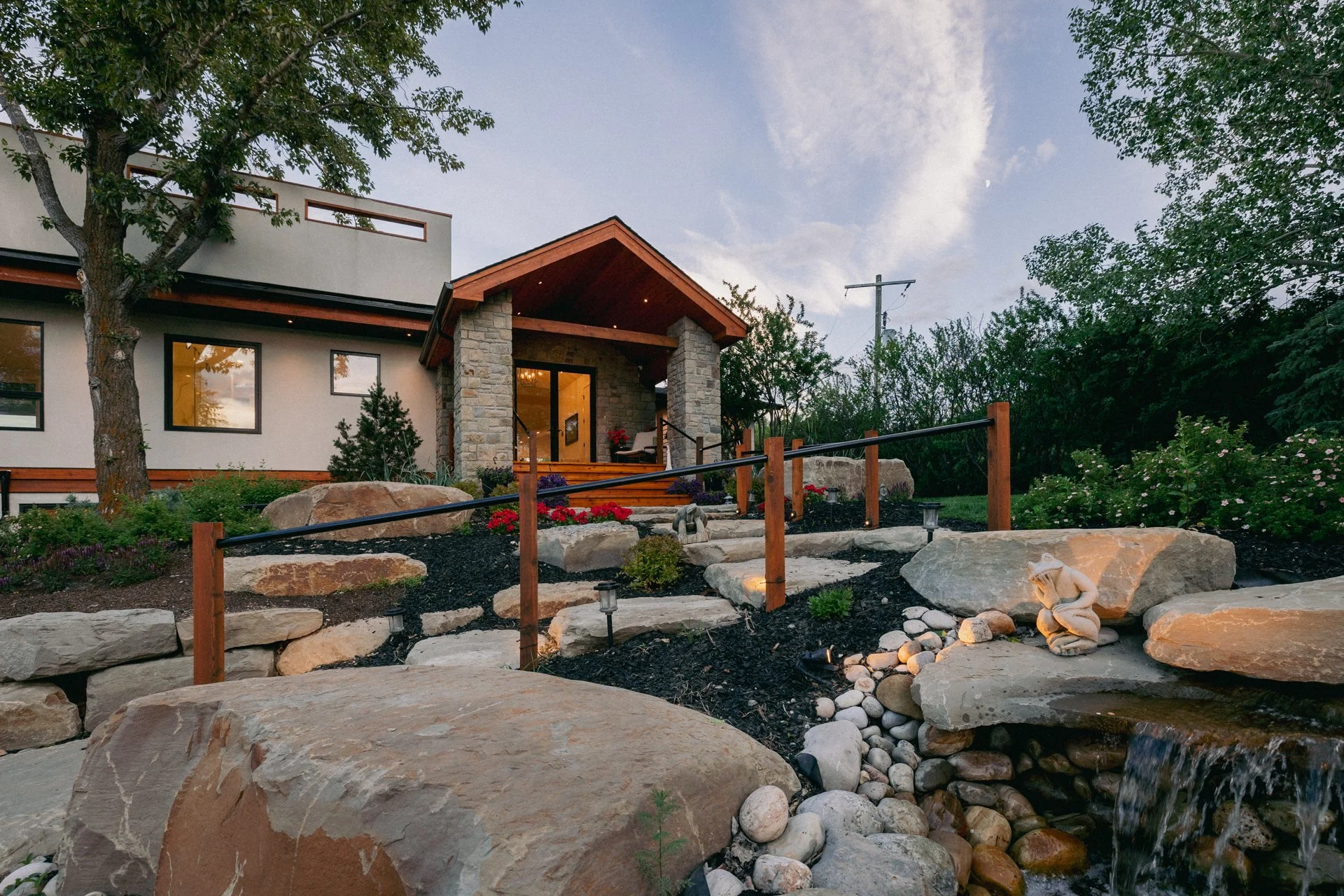 A modern house with stone and wood exterior, surrounded by landscaped garden with rocks and plants, featuring a small waterfall and pathways, during sunset with clear sky.