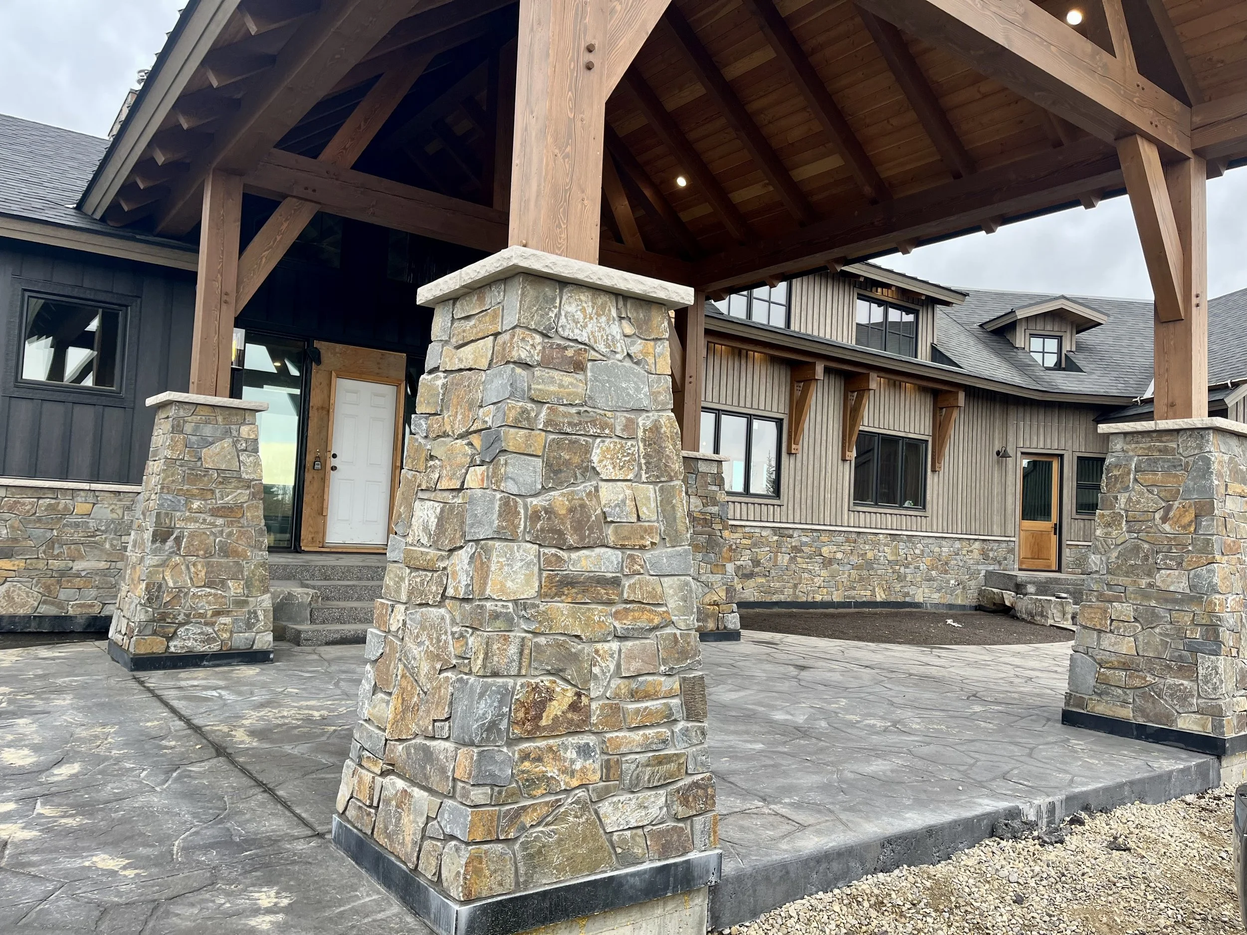Front porch of a modern house with stone pillars, a wooden roof structure, and a concrete patio.