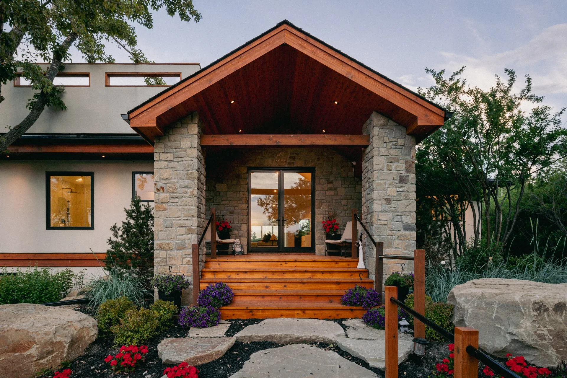 Front porch of a modern house with wooden stairs leading to glass double doors. The porch is supported by stone columns and decorated with potted plants and flowers. Large rocks and plants are in the garden area surrounding the steps.