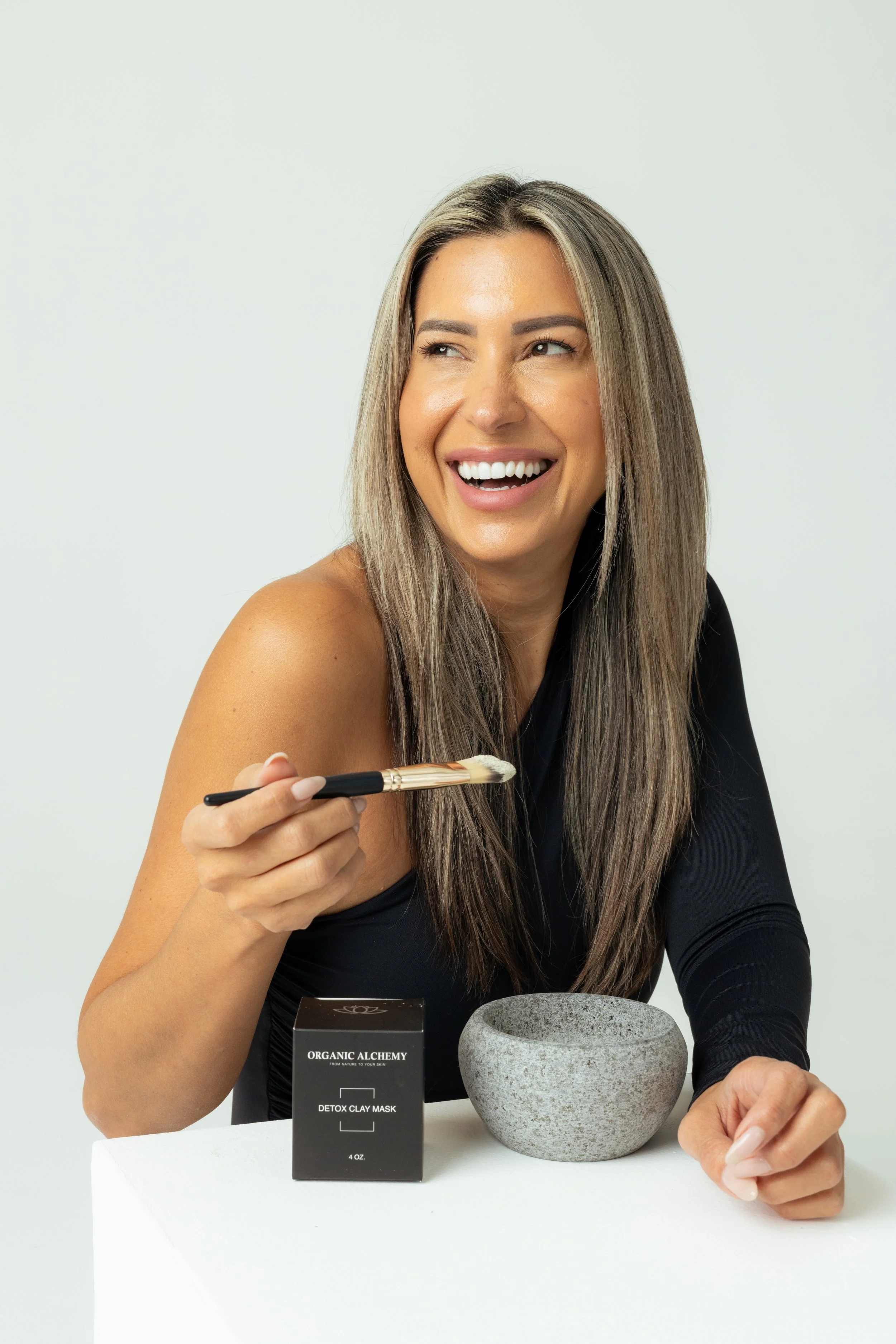 A woman smiling and holding a makeup brush near her shoulder, sitting at a white surface with a gray stone bowl and a black box labeled 'Organic Alchemy Detox Clay Mask' in front of her.