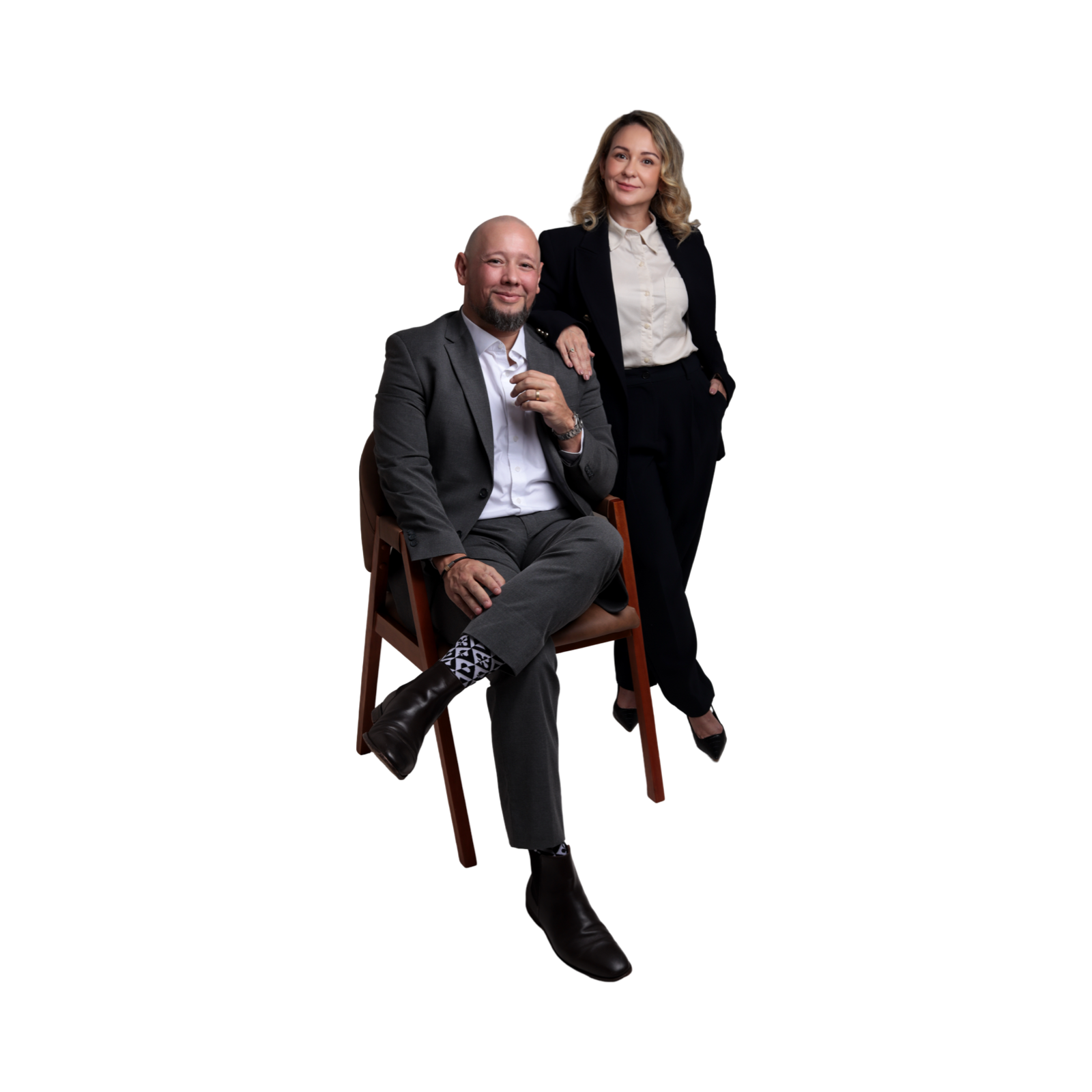 Homem vestindo terno cinza sentado em cadeira, mulher com terno preto ao lado, ambos com expressão confiante, fundo escuro.