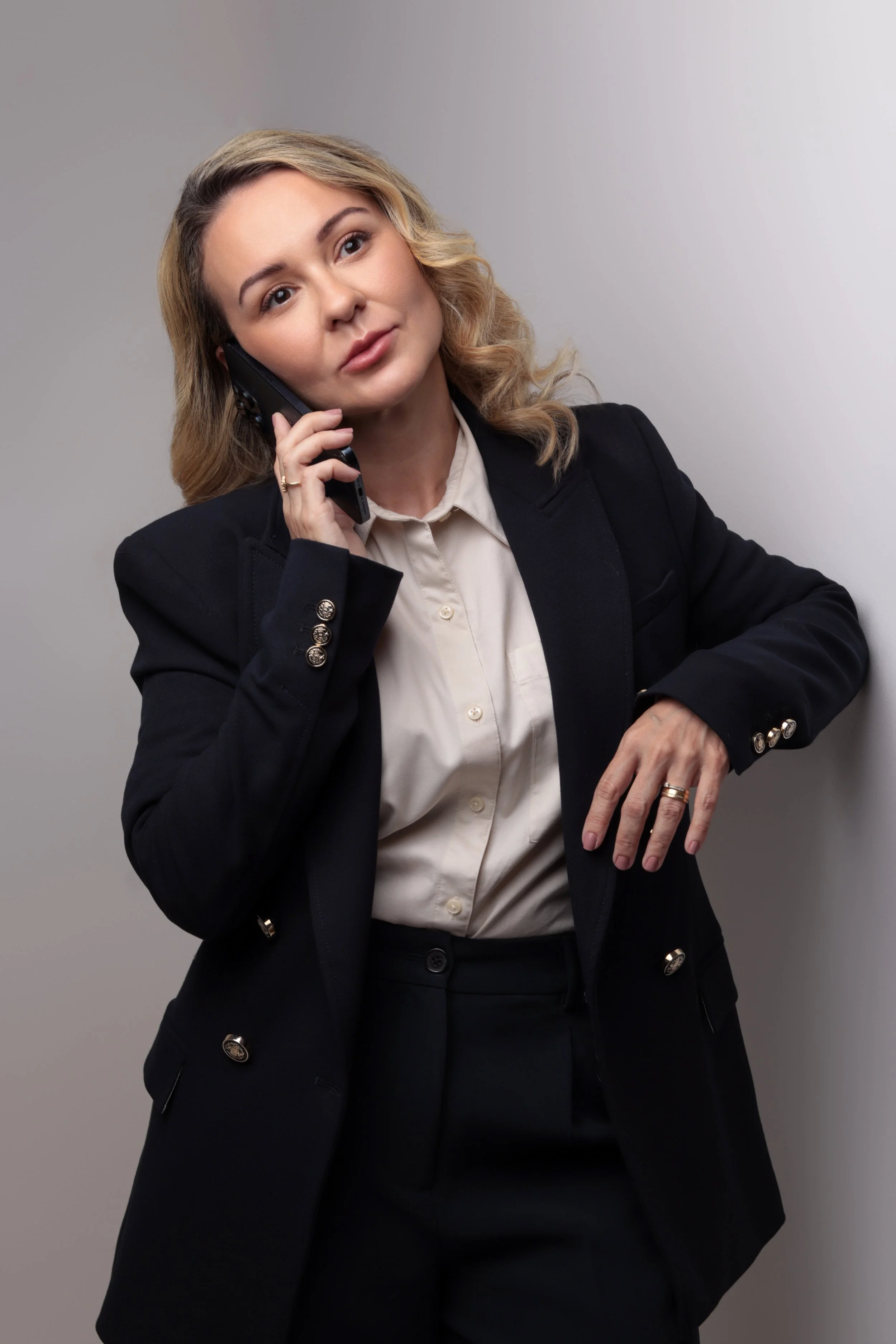 Mulher de cabelos cacheados ao telefone, vestida com blazer preto e blusa clara, posando junto a uma parede cinza.