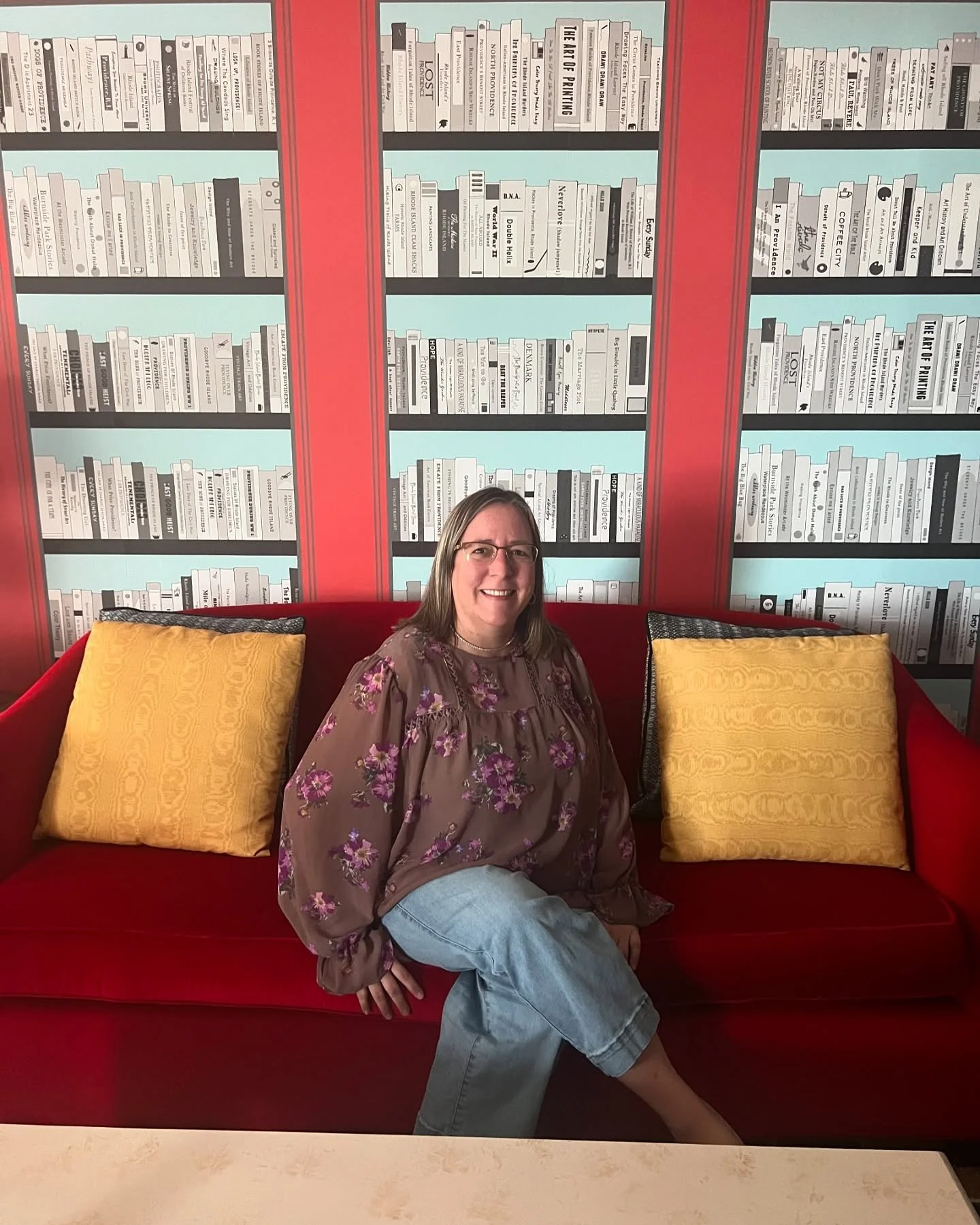 A woman sitting on a red sofa with yellow pillows, in front of a bookshelf mural on the wall.