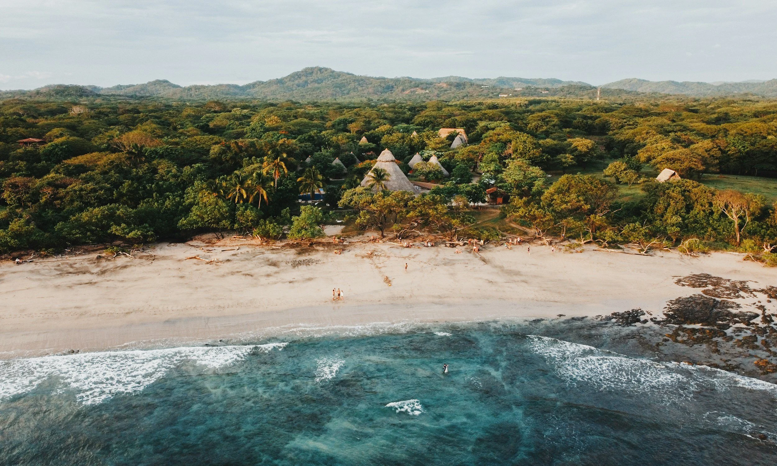 Aerial view of a tropical beach with white sand, turquoise water, and green trees, with thatched-roof structures nestled among the trees and mountains in the background.