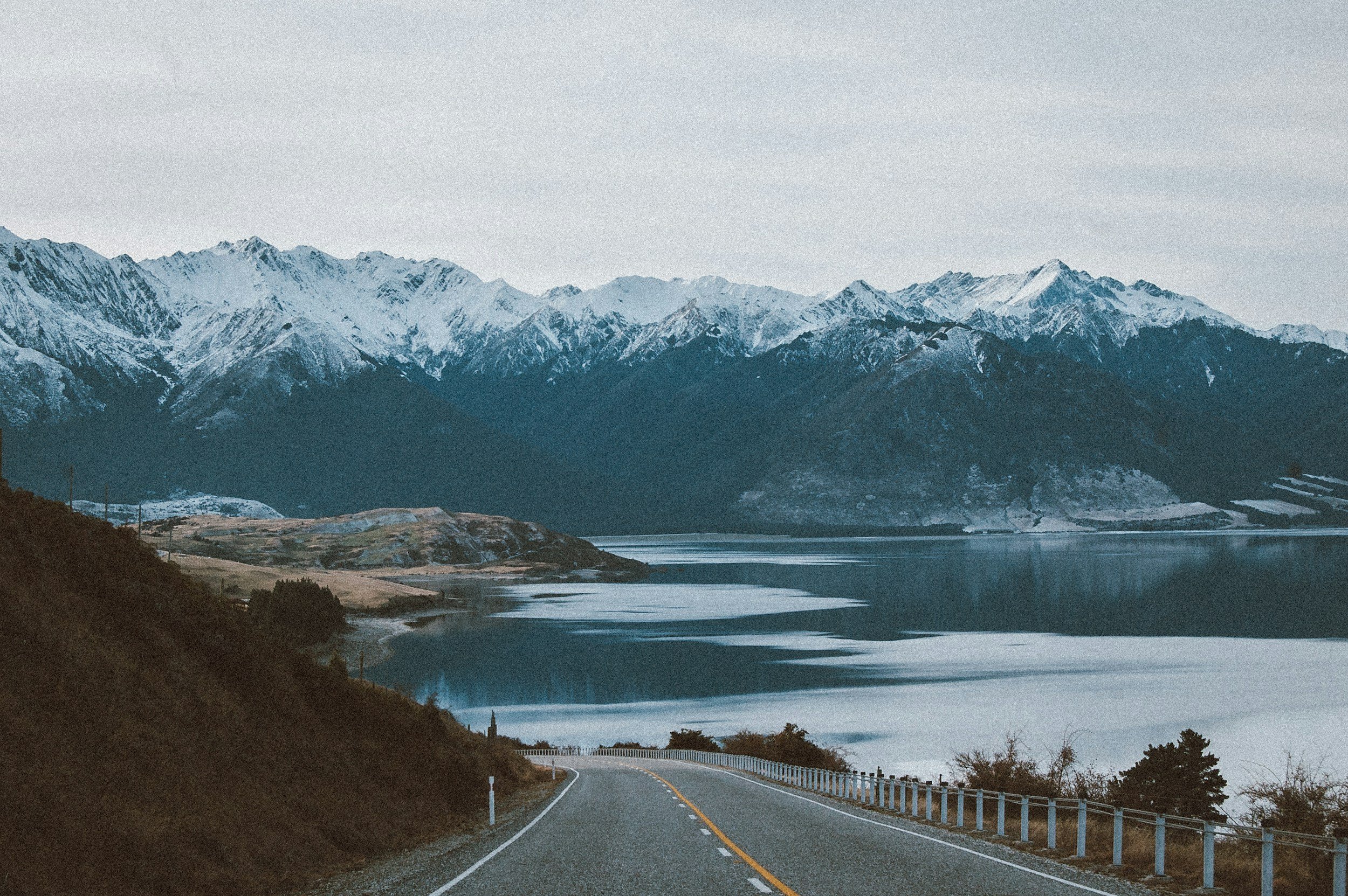 A winding road leading to a lake with snow-capped mountains in the background under an overcast sky.