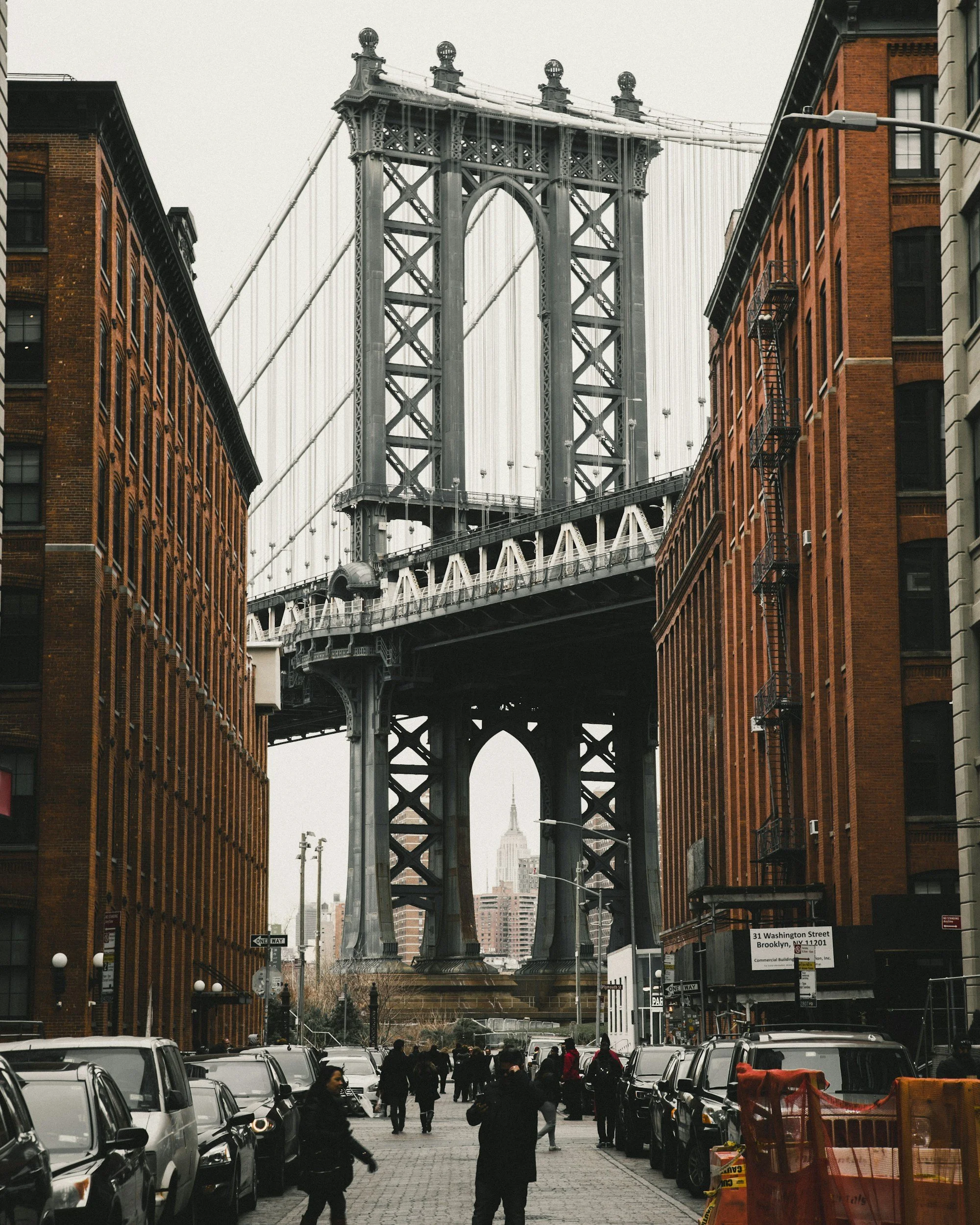 View of the Brooklyn Bridge in New York City, with people walking and cars parked along the street in the foreground and the Empire State Building visible in the background.