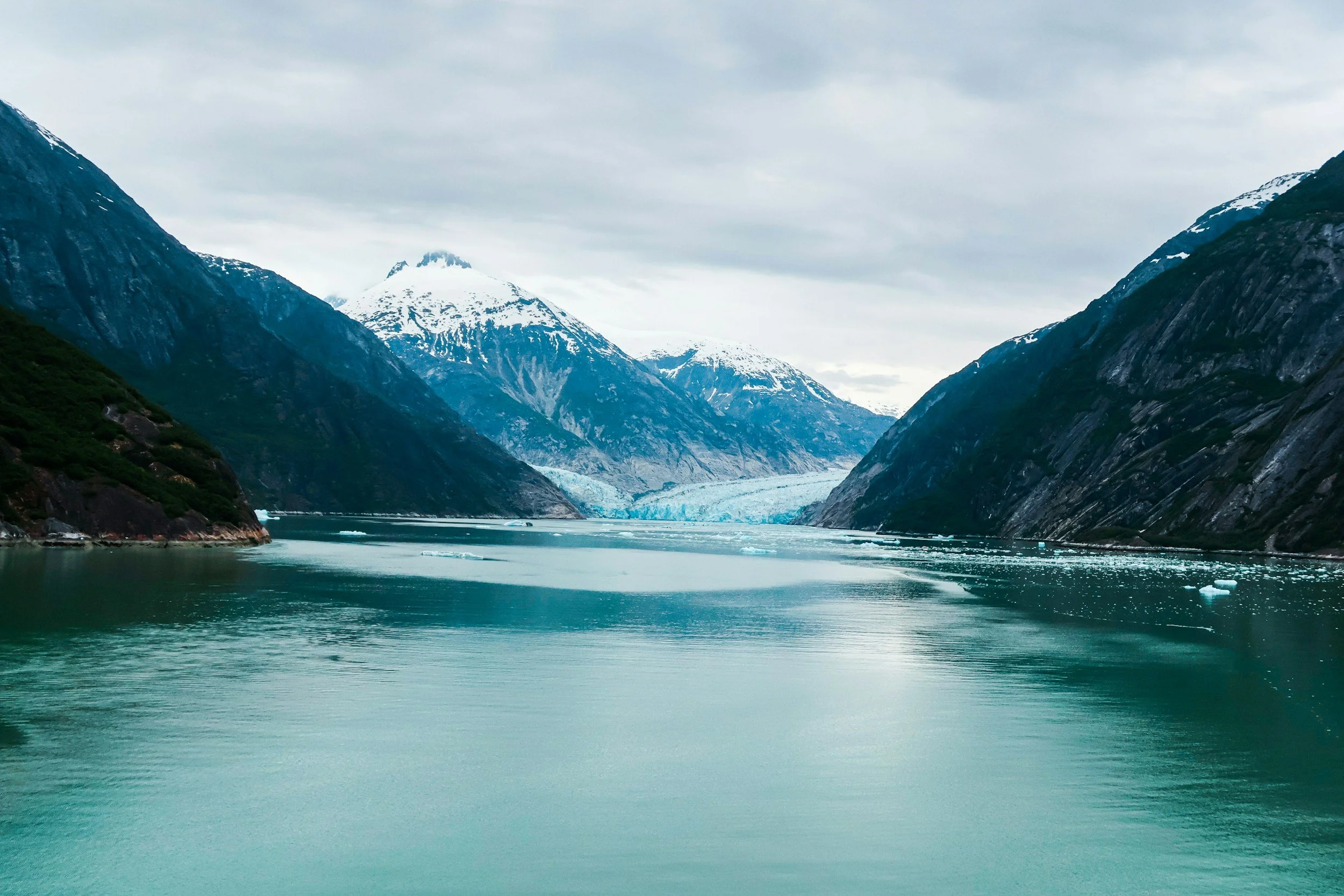 Scenic view of a glacier-fueled fjord surrounded by snow-capped mountains under an overcast sky.
