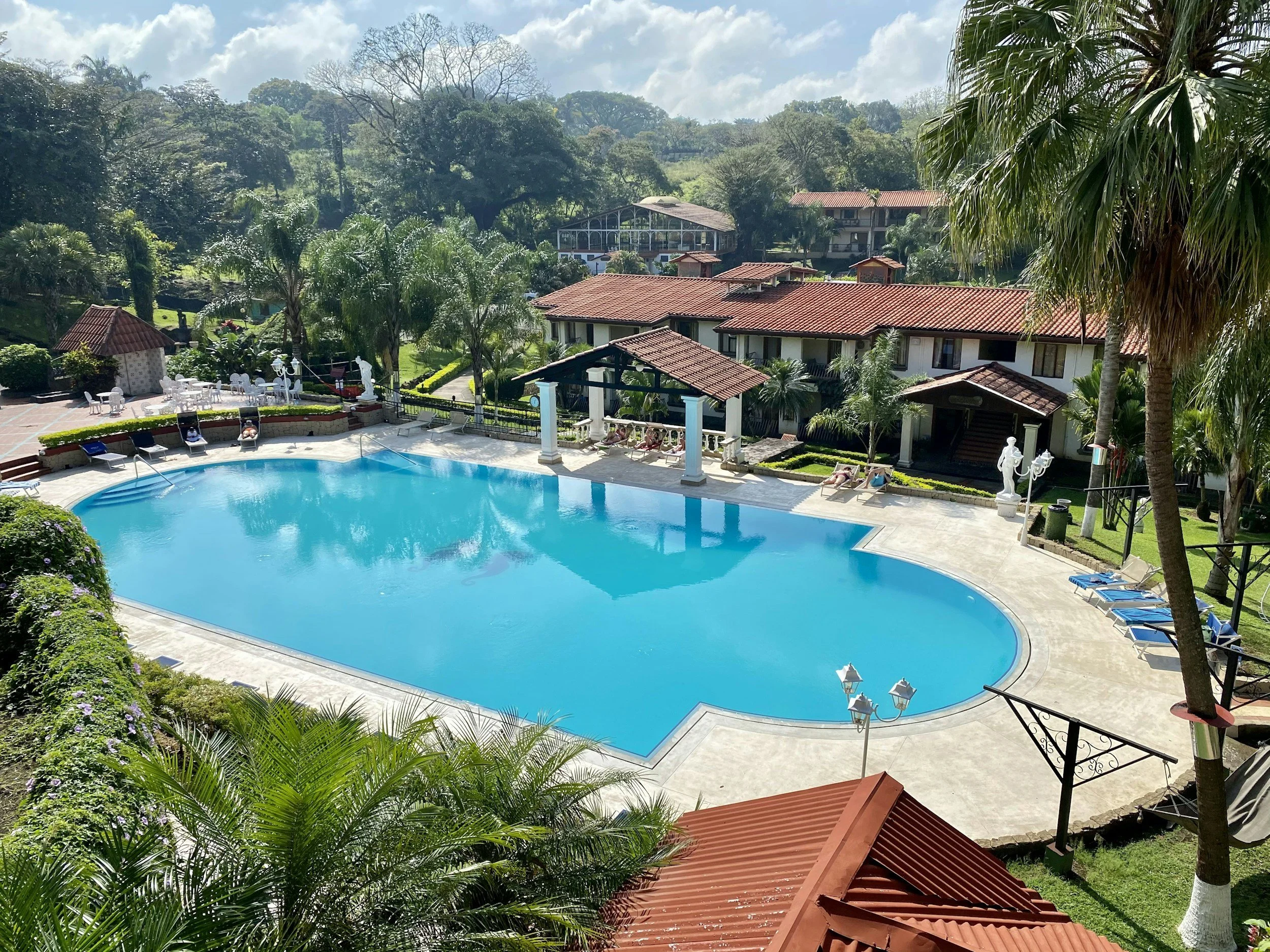 A large outdoor swimming pool surrounded by lounge chairs and lush greenery, with a resort building in the background and a partly cloudy sky above.